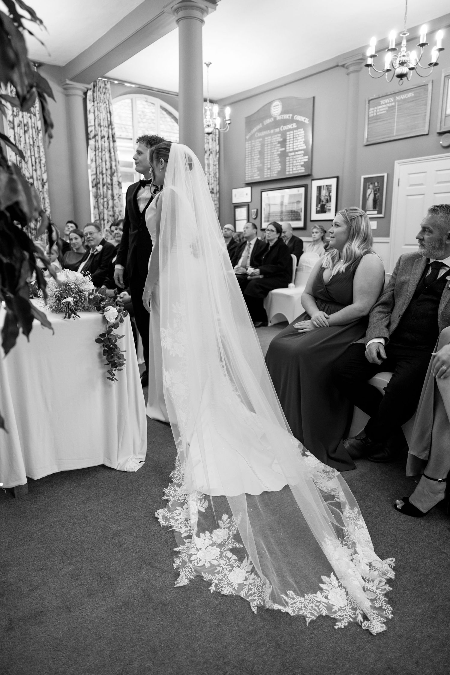 A wedding celebration in an elegant room with guests admiring the bride's flowing dress and veil.