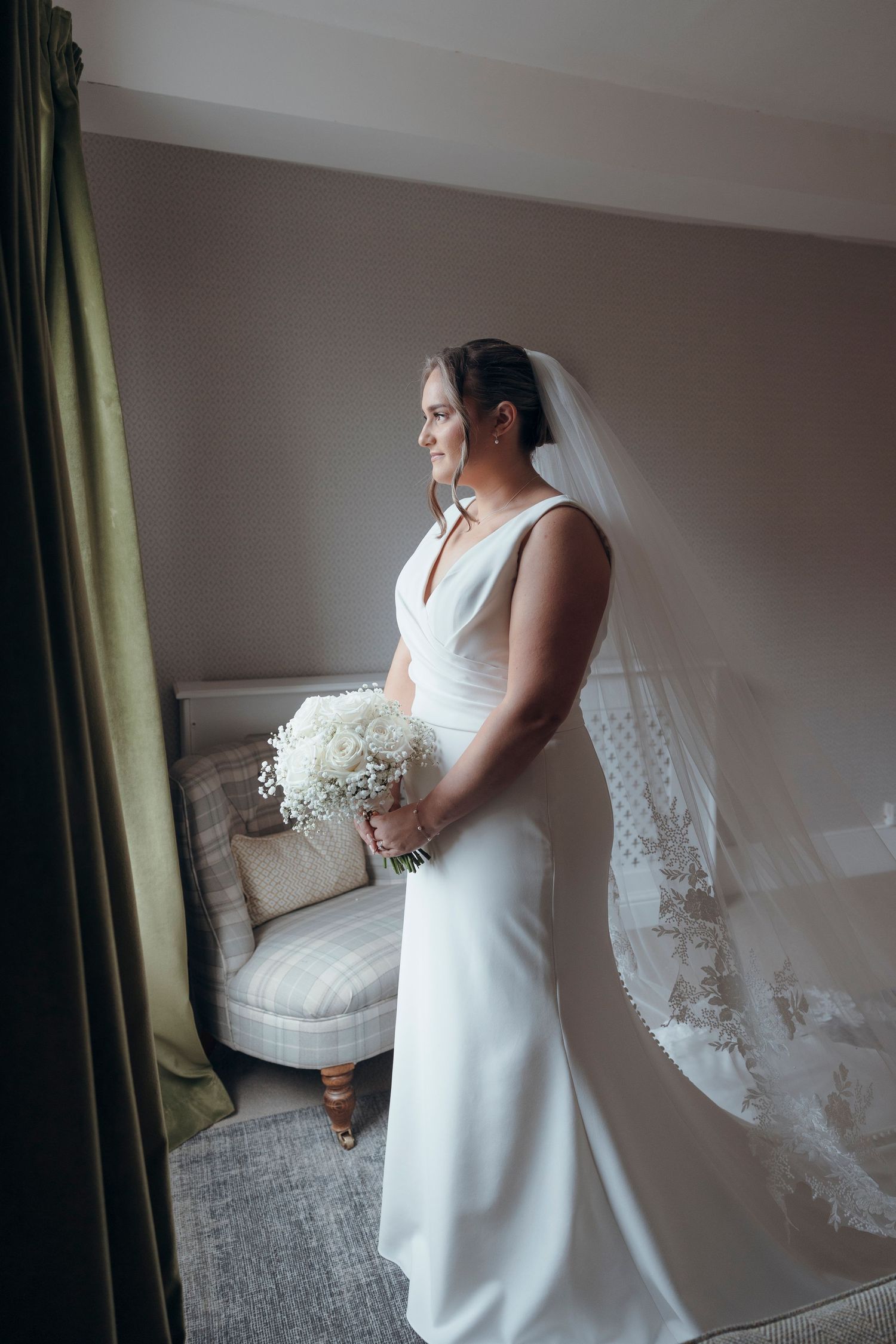 A bride in an elegant white dress holding a small bouquet near a window.