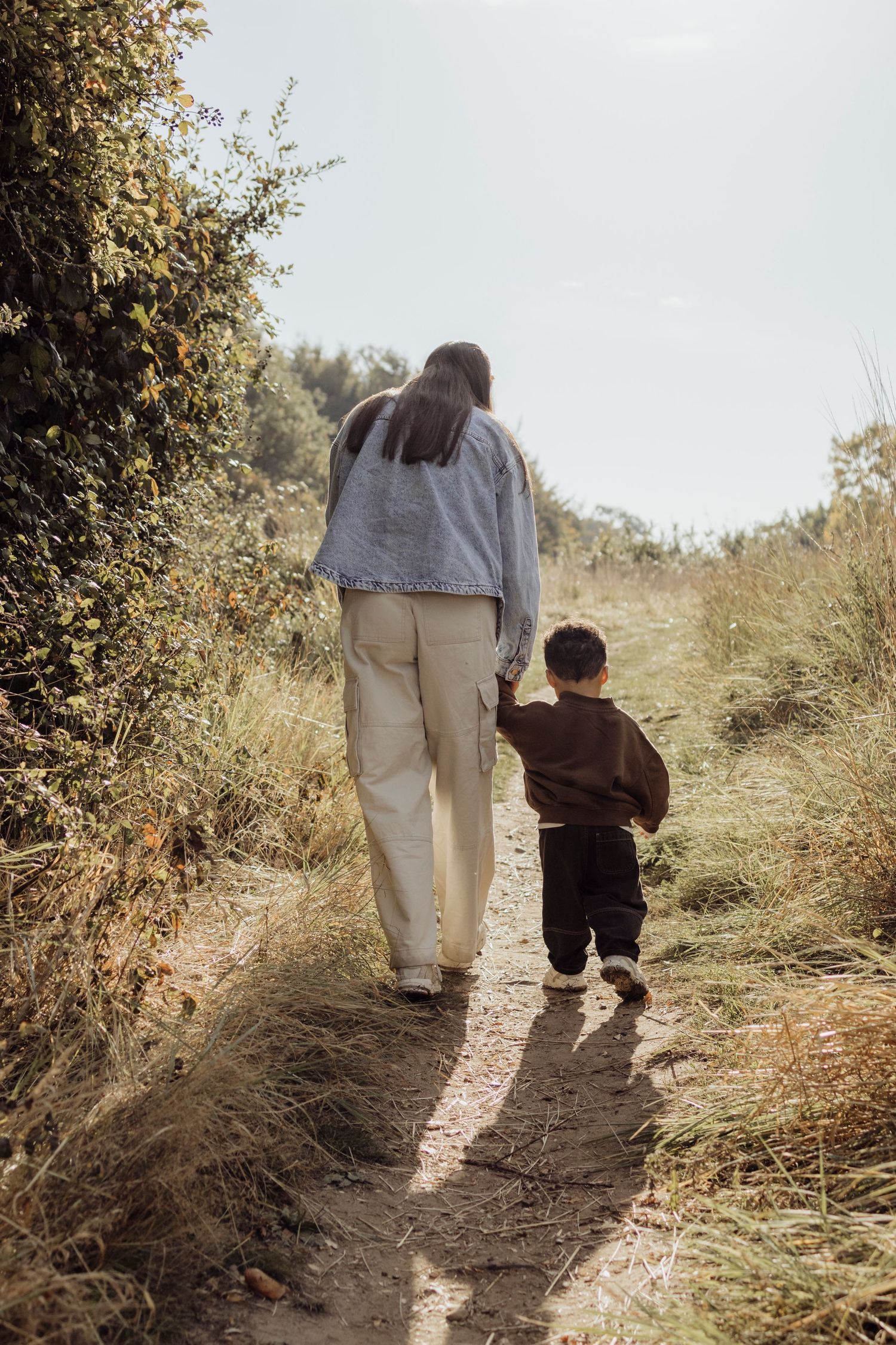 Two figures walk together on a winding dirt path through tall grass and autumn foliage on a sunny day.