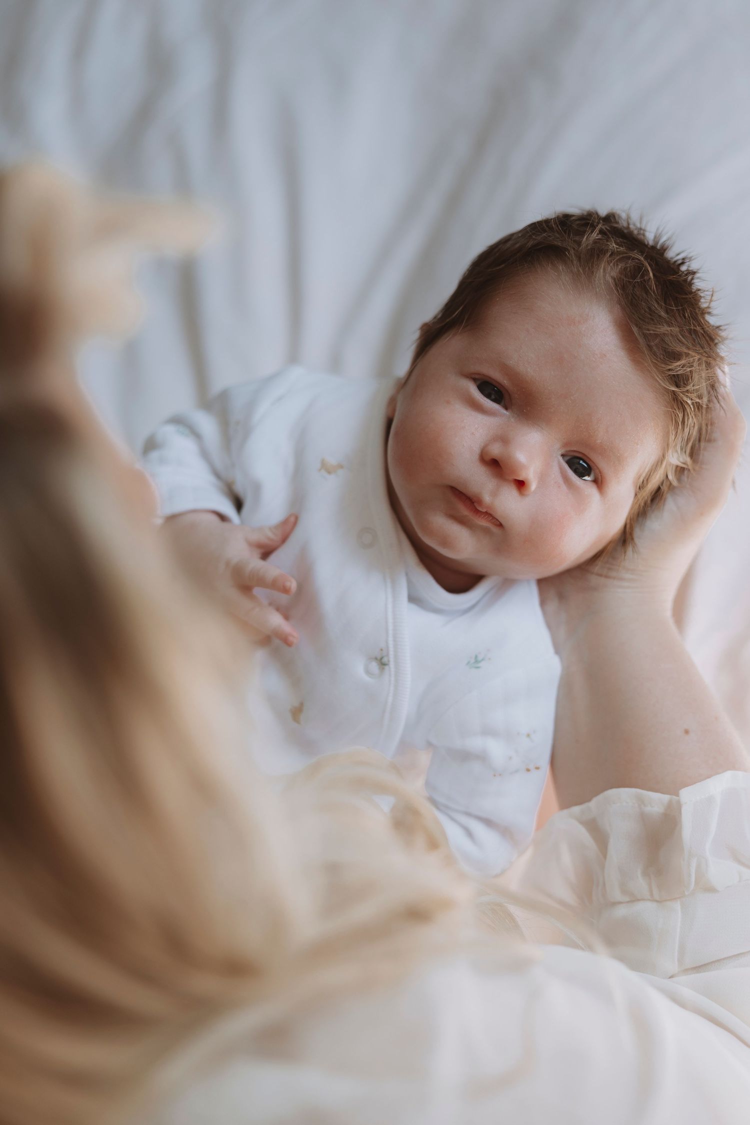 A newborn baby in a white onesie lying on white bedding, gazing up at a blonde woman leaning over them.