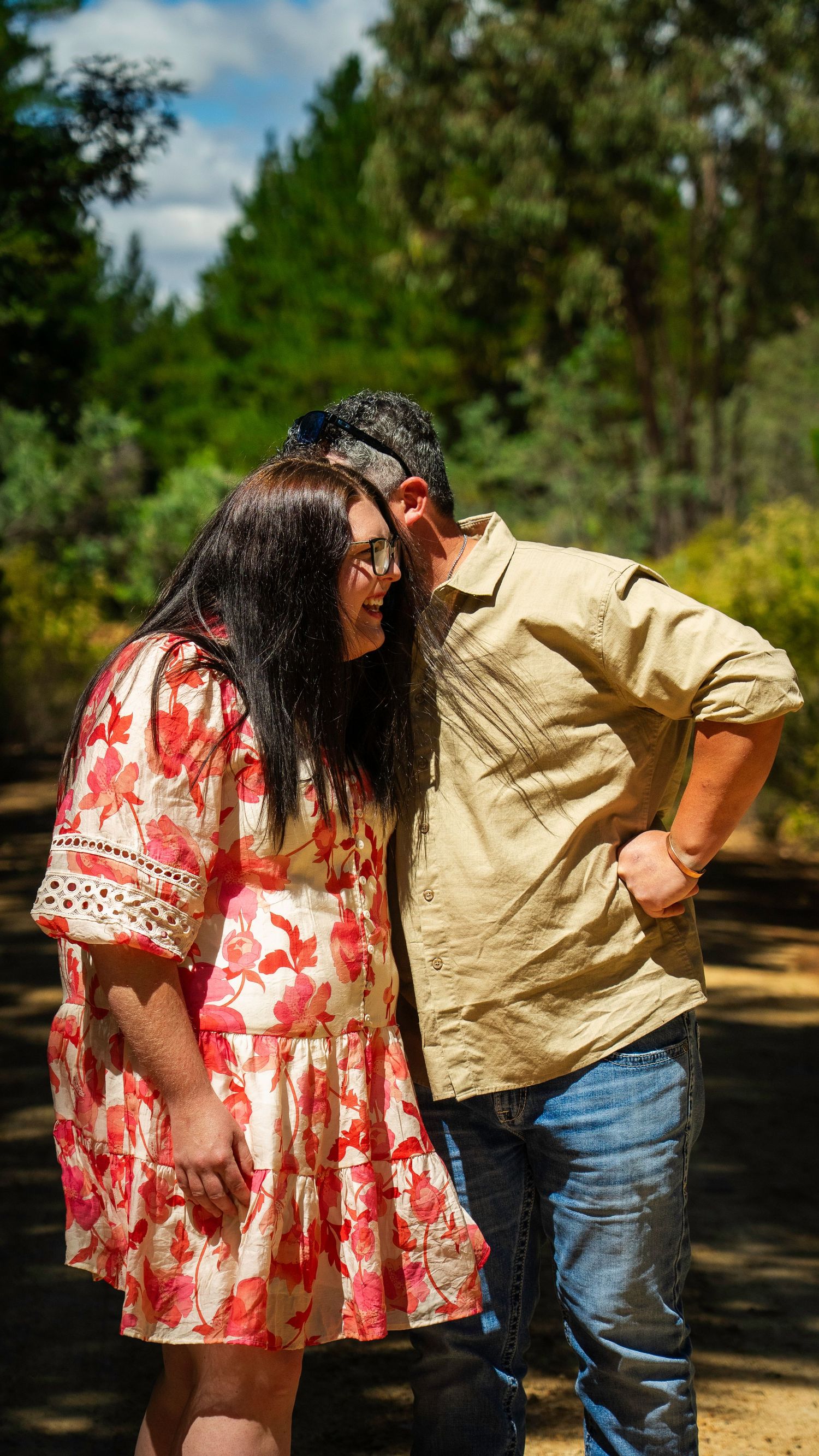 Couple sharing a tender moment on a forest path, woman in floral dress and man in beige shirt embracing outdoors.
