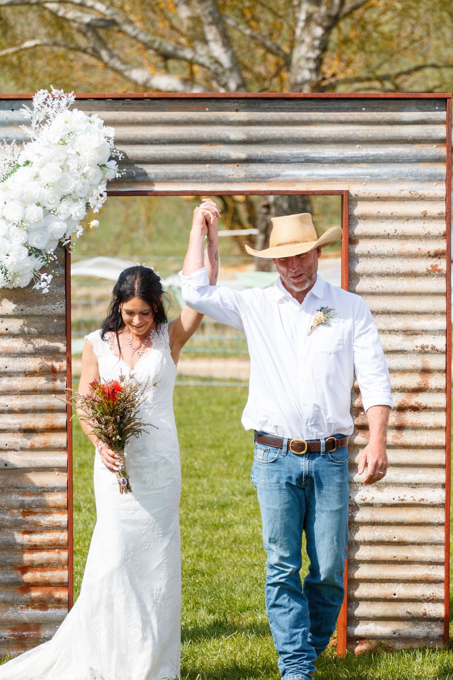 Bride and groom exit rustic corrugated metal arch, groom in cowboy hat raises bride's hand in celebration.