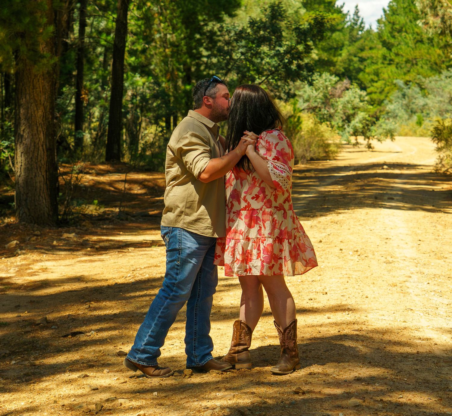 Couple dancing and kissing on a sunny dirt path surrounded by tall trees in a forest setting.