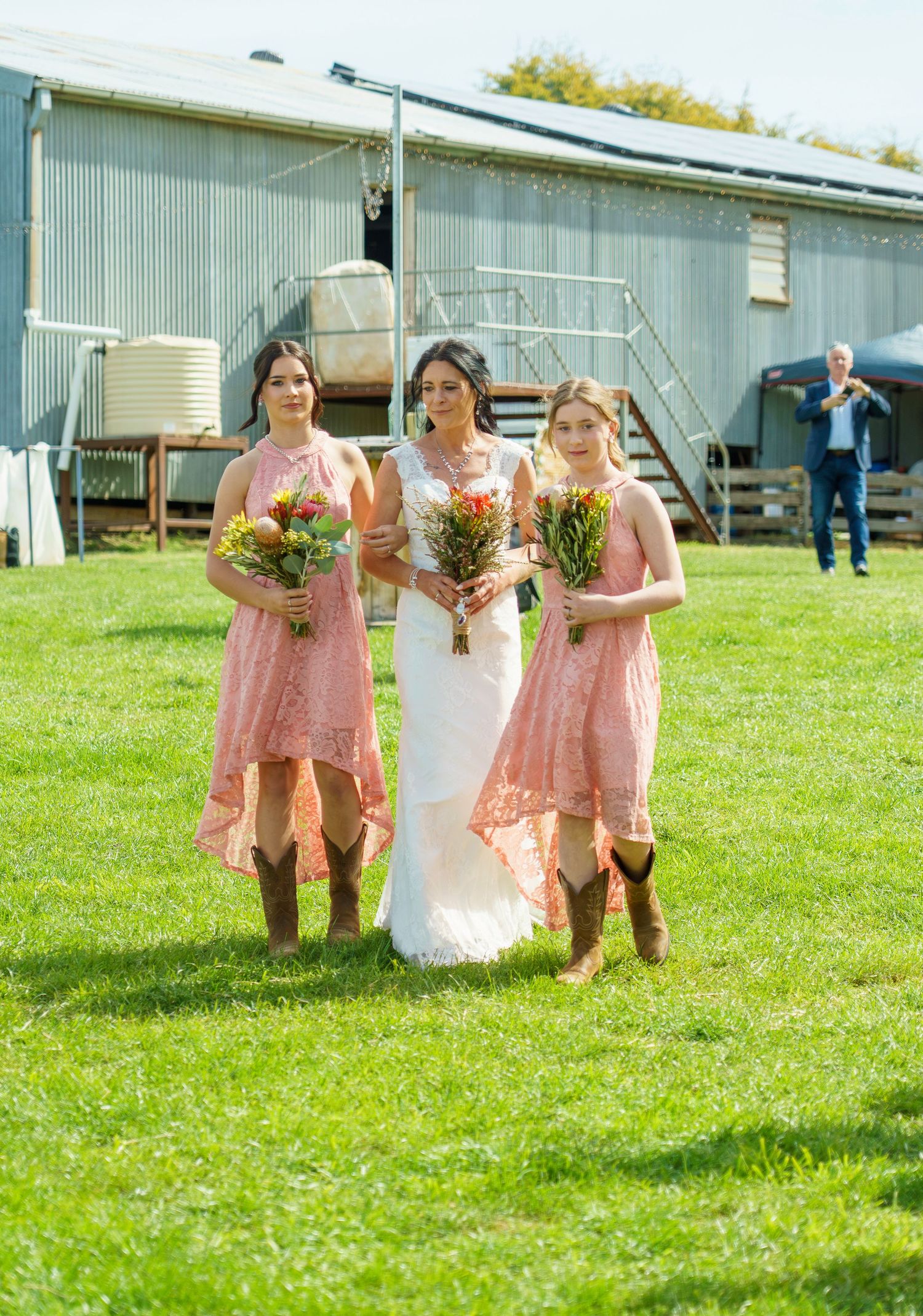 Bride in white gown walks with two bridesmaids in pink lace dresses and cowboy boots holding wildflower bouquets.