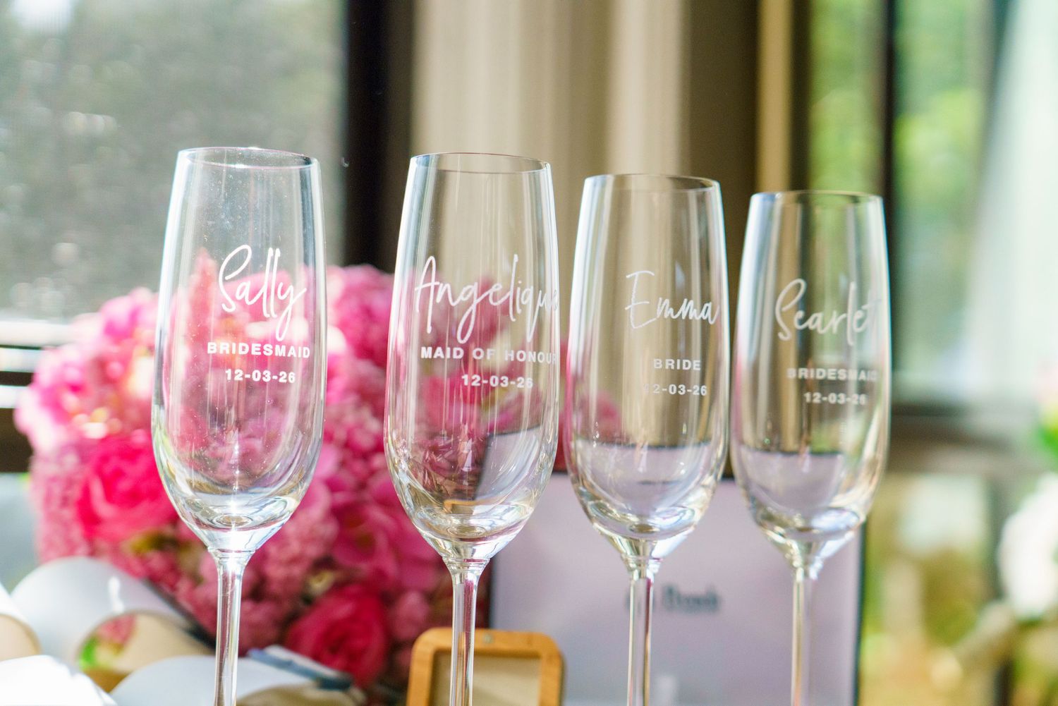 Four personalized champagne flutes with white calligraphy names for bridesmaids displayed in front of pink floral arrangements.