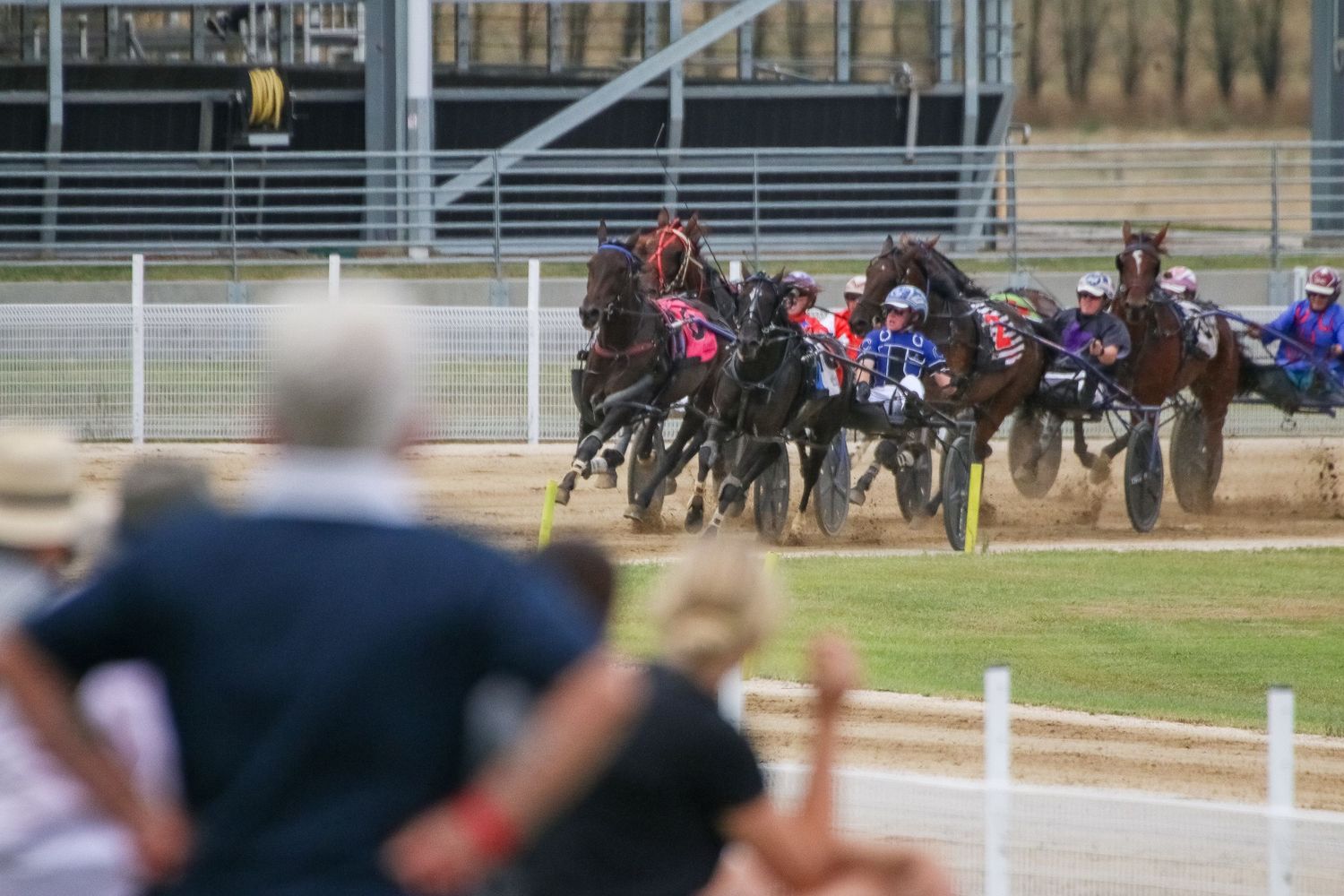 Spectators watch harness racing competition from behind track barriers.