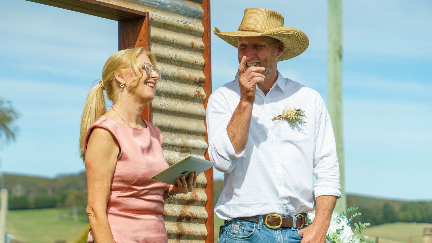 A woman and man in a cowboy hat chat outdoors near a rustic corrugated iron shed on a sunny farm day.
