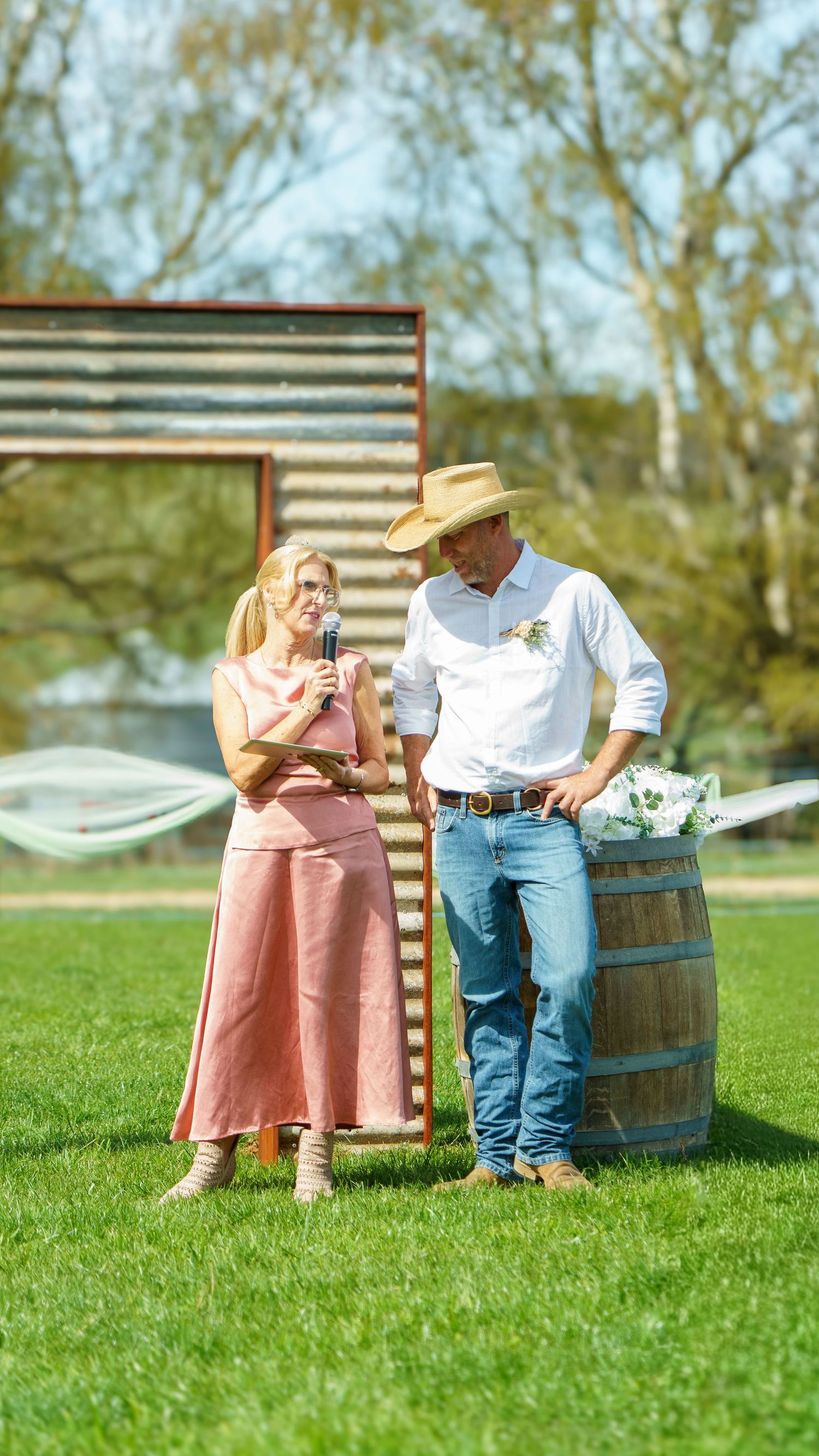 Couple in rustic outdoor setting, woman in pink dress speaking into mic, man in cowboy hat leaning on wooden barrel.