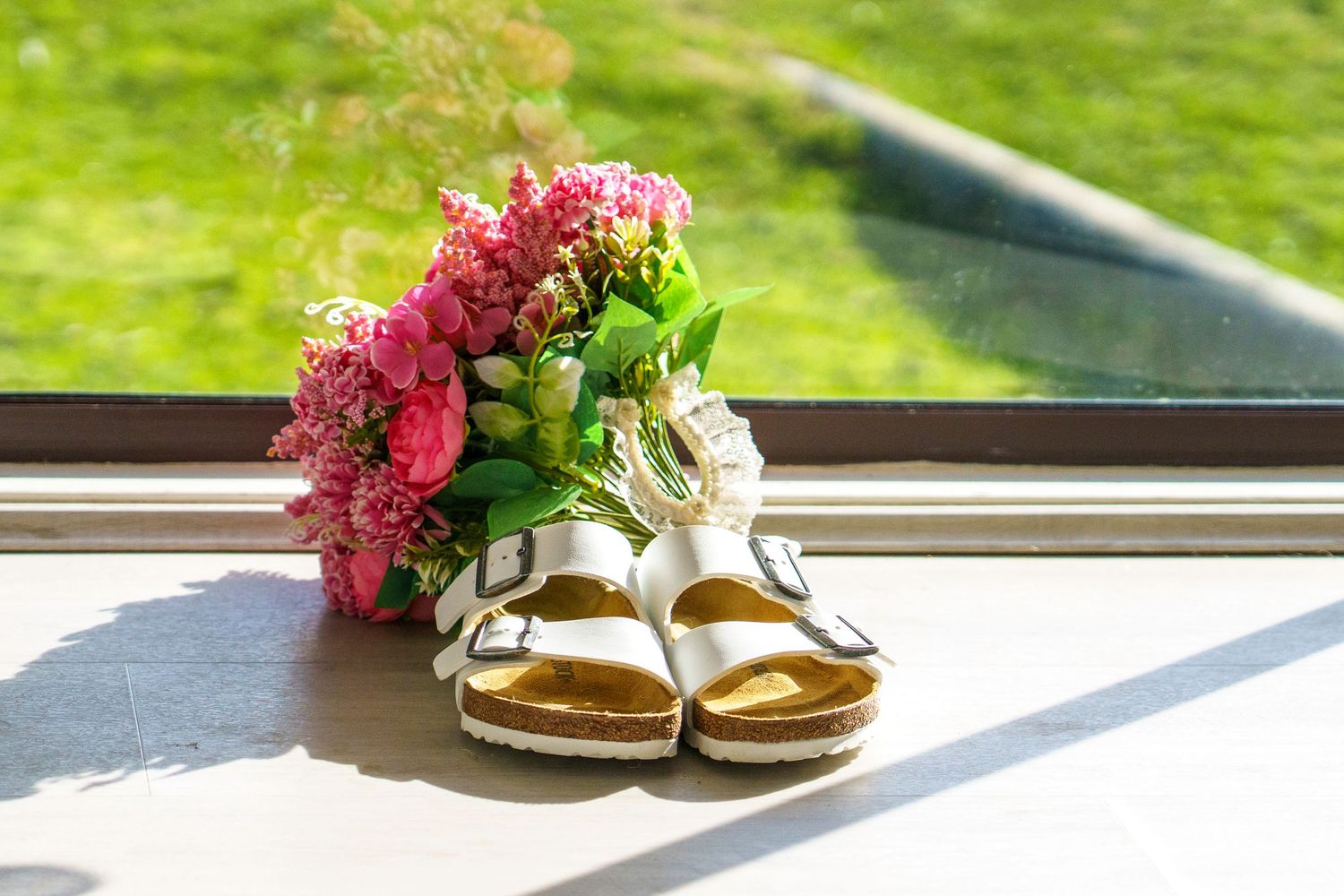 White Birkenstock sandals beside a pink floral bouquet on a sunny windowsill with green grass in background.