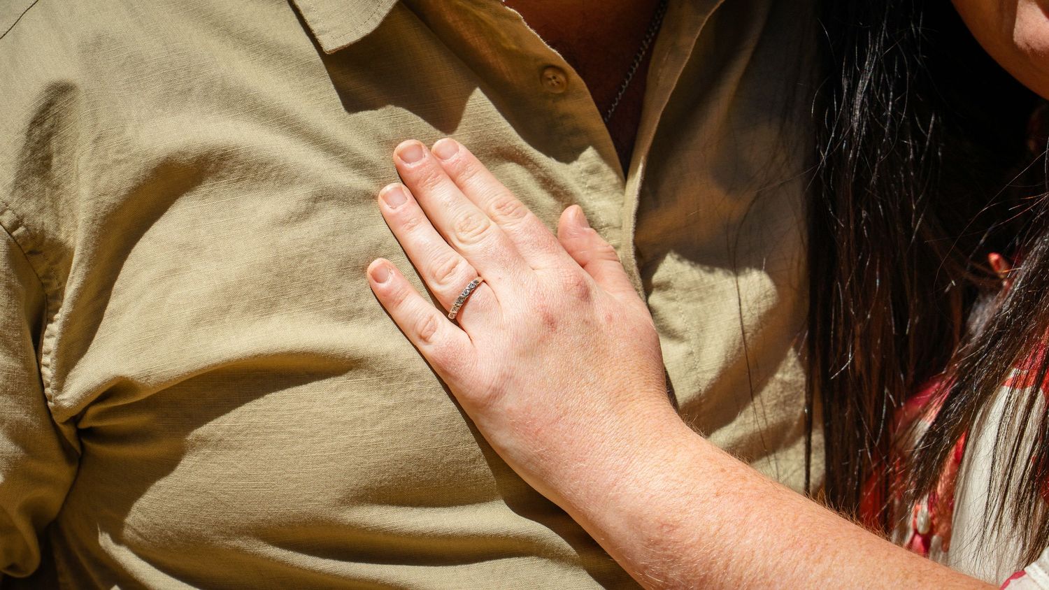 Close-up of a woman's hand with an engagement ring resting on a man's chest outdoors.