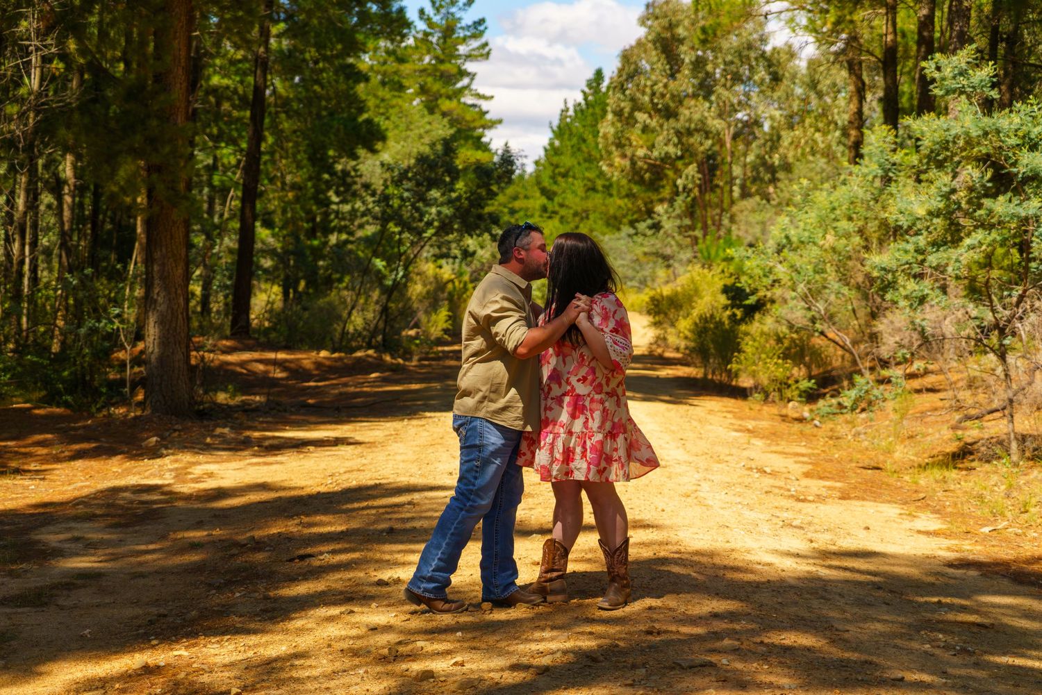 Couple sharing a romantic kiss on a sunlit forest dirt path surrounded by tall pine trees.