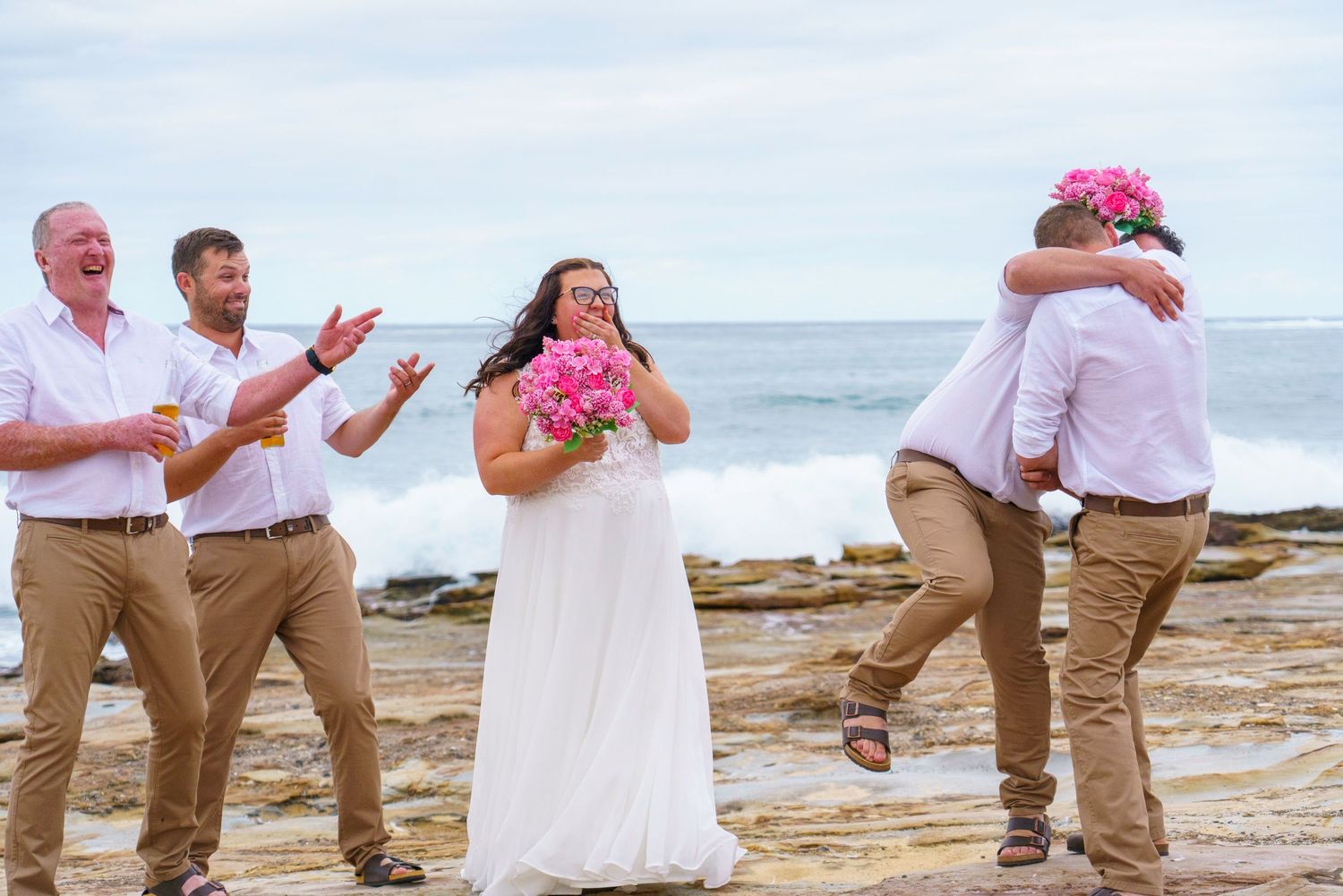 Beach wedding celebration with bride in white dress holding pink bouquet, groom kissing guest, groomsmen cheering by ocean.