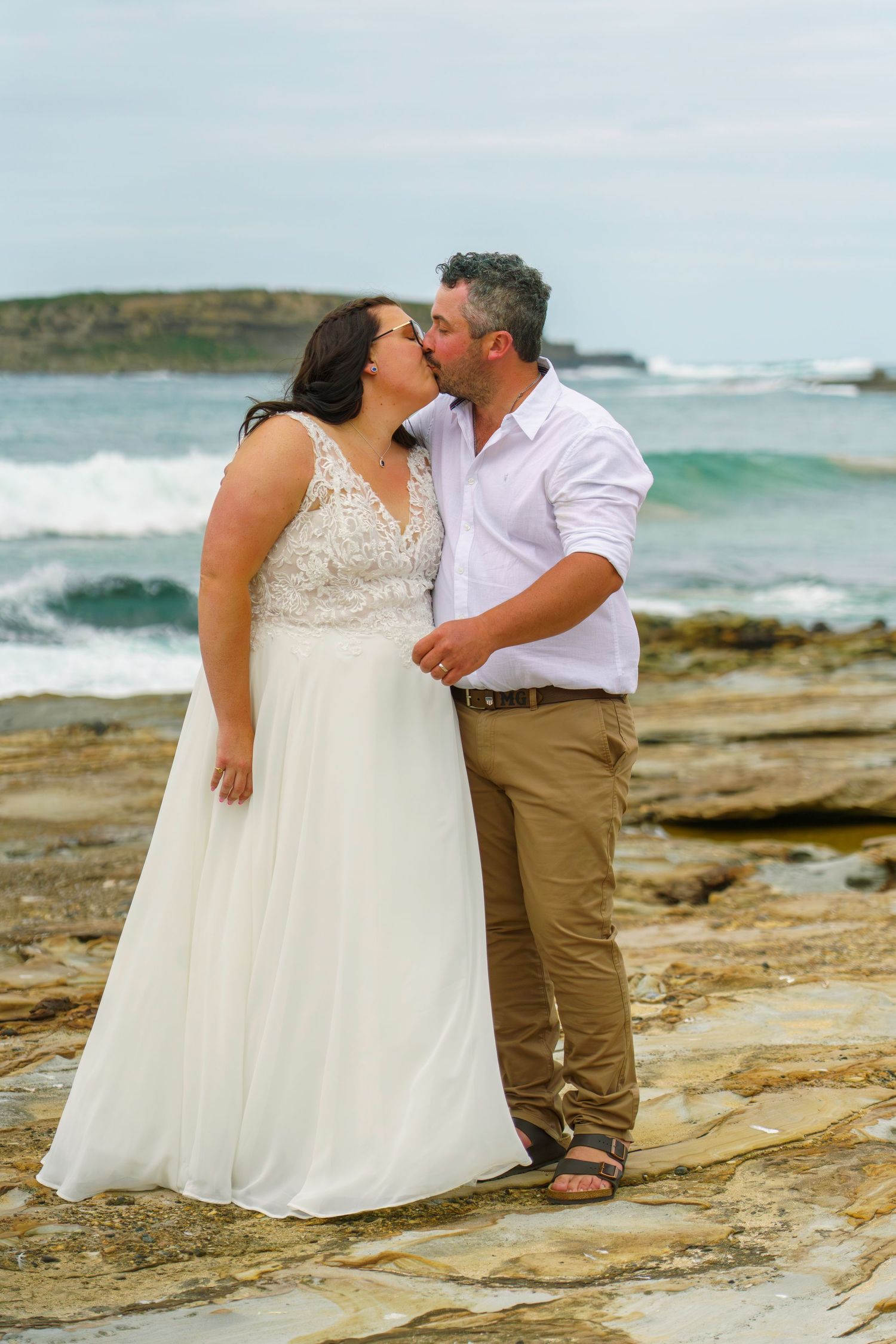 Bride and groom kissing on rocky coastal shoreline, bride in white lace gown, ocean waves in background.
