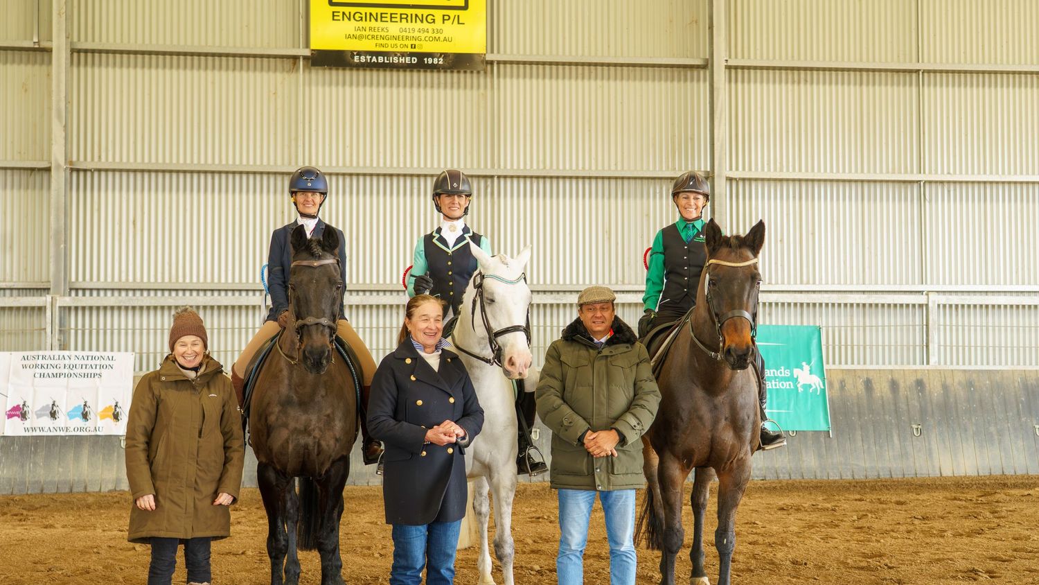 Multiple riders and horses line up along the wall of an indoor equestrian facility.