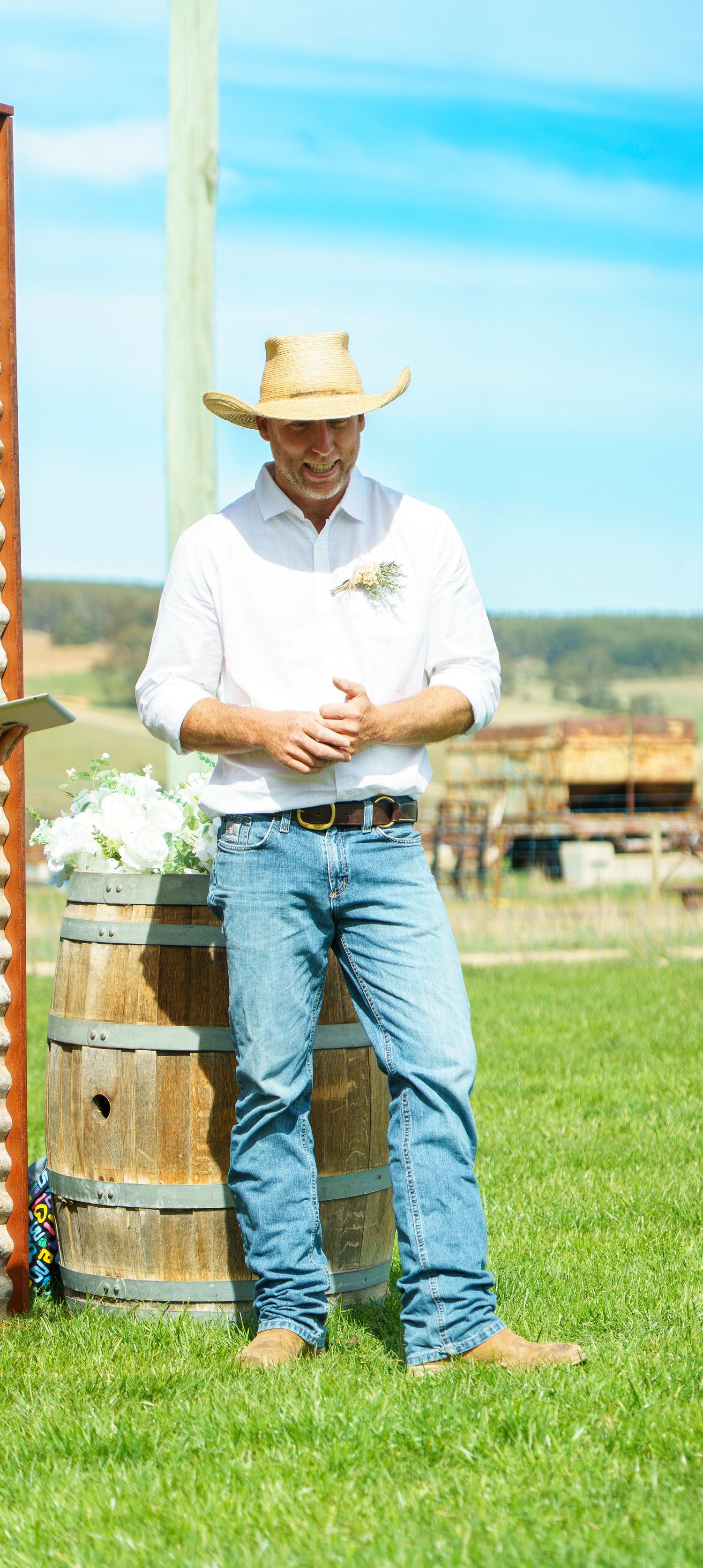 Smiling groom in cowboy hat and white shirt standing by rustic wooden barrel at outdoor wedding.