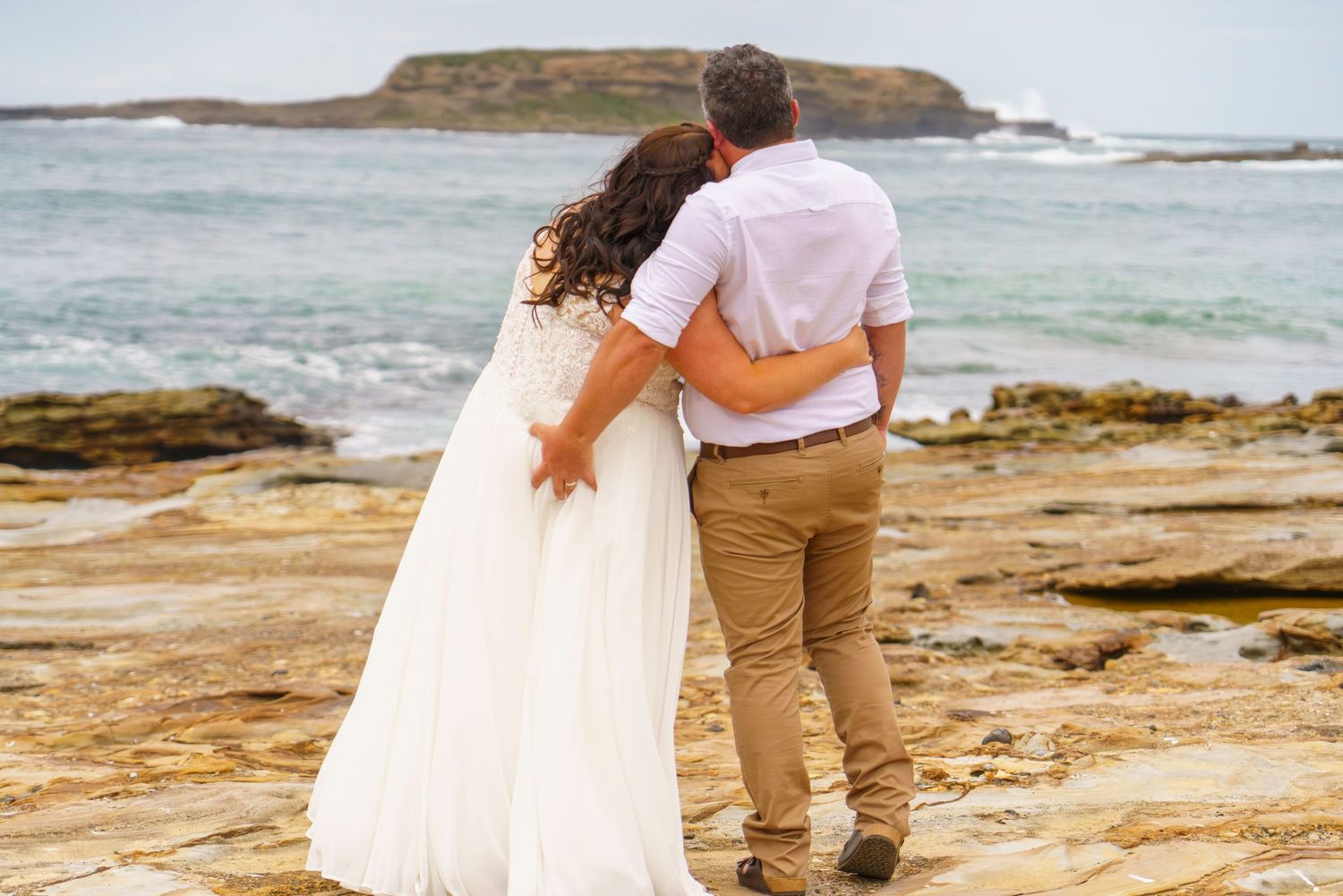 A couple in wedding attire embraces on rocky coastal cliffs, gazing at turquoise ocean waves under an overcast sky.