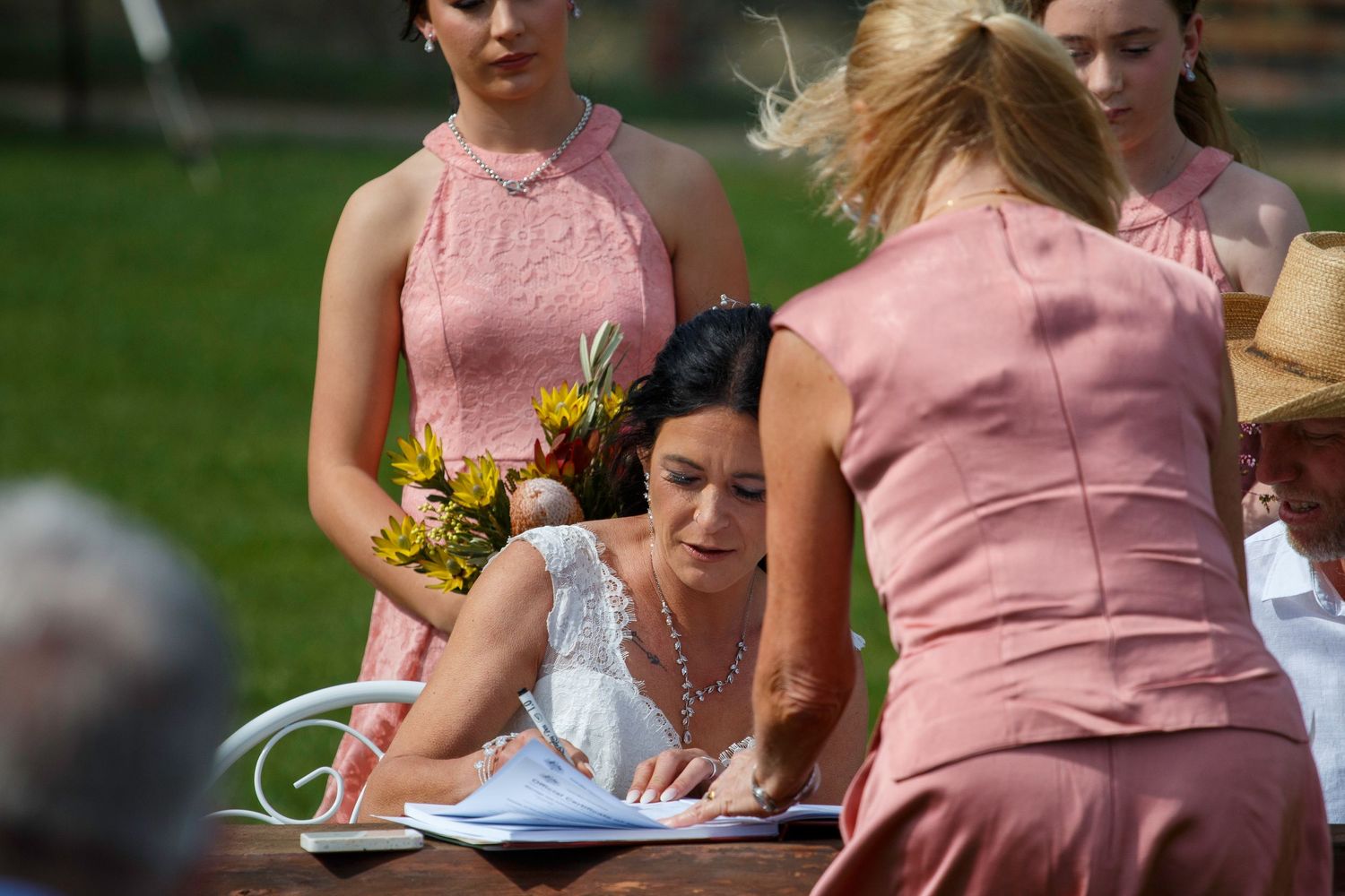 Bride in white dress signs wedding certificate outdoors surrounded by bridesmaids in pink dresses.
