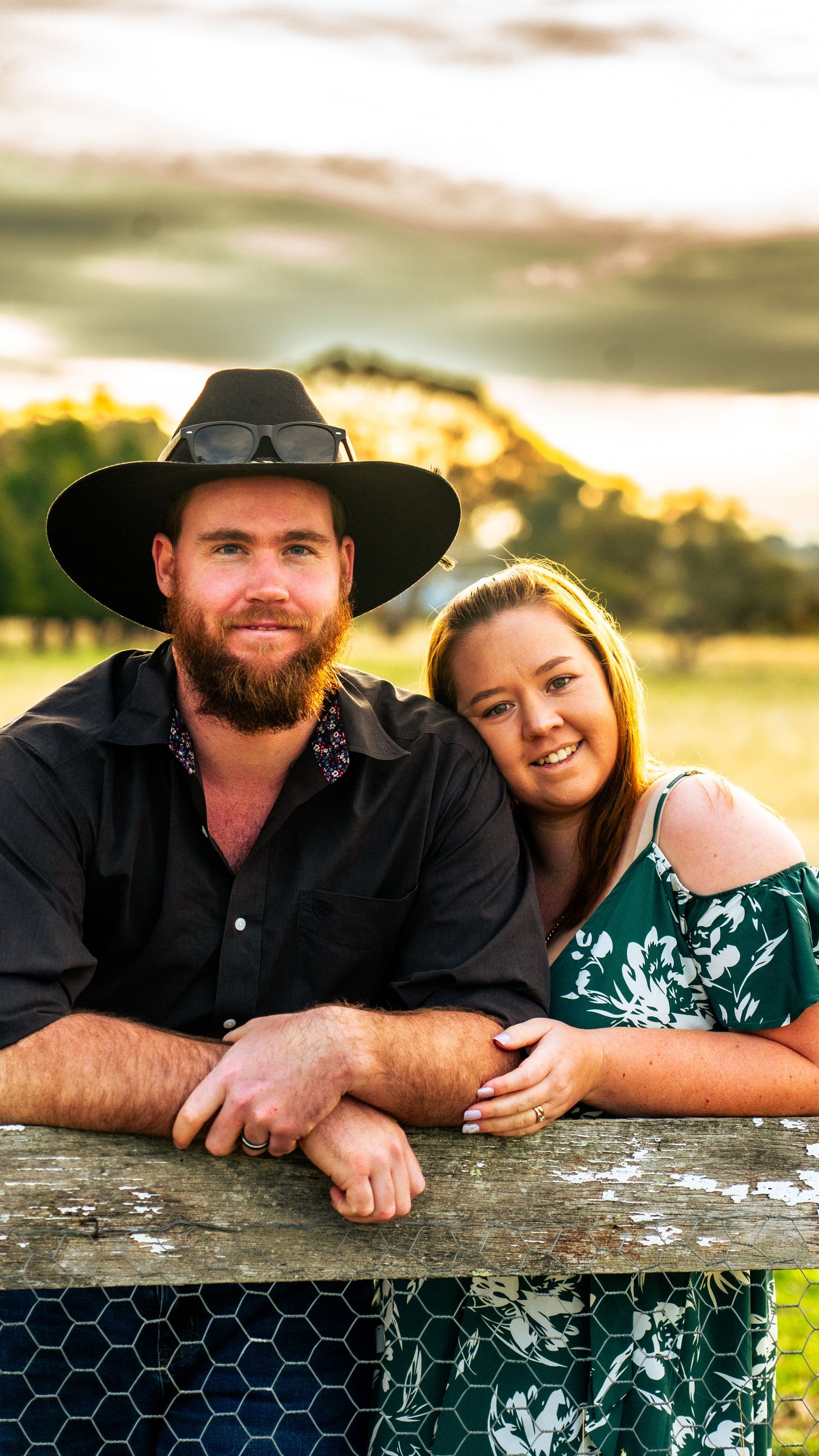 Couple poses together by rustic fence during golden hour photo session.