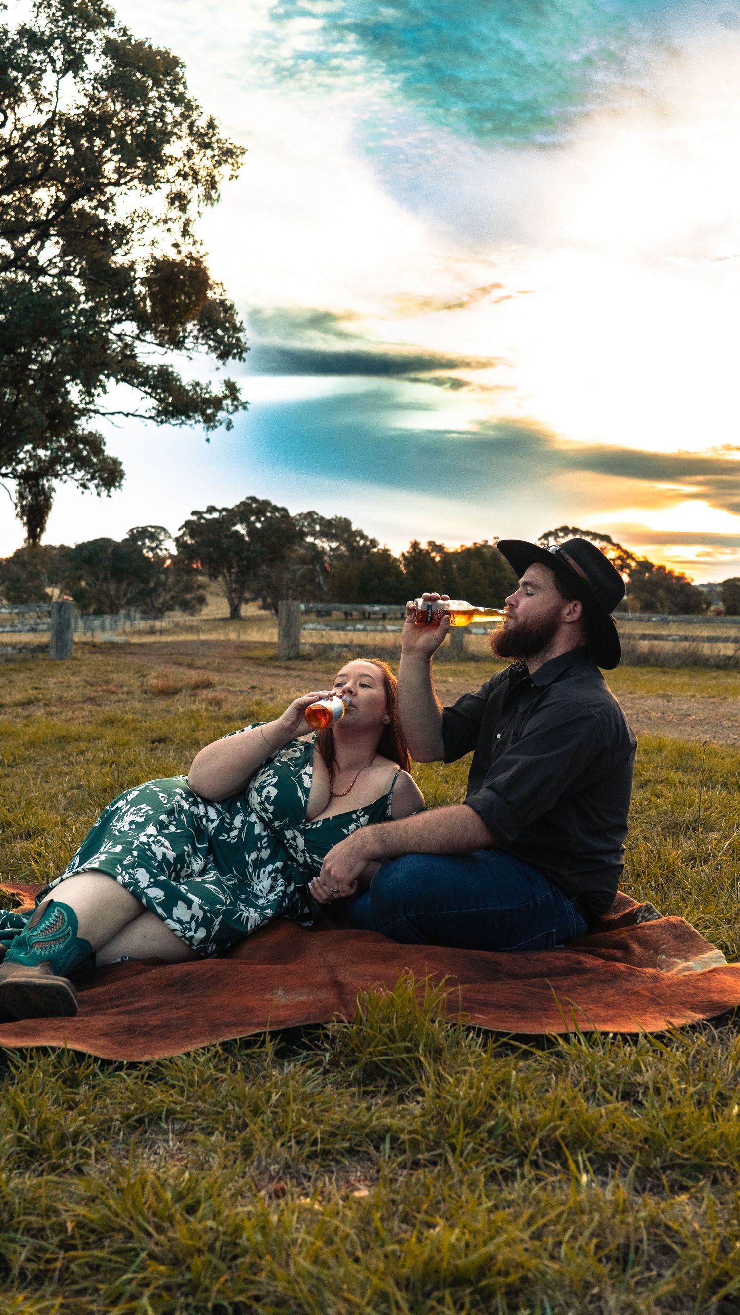 Couple relaxes on blanket drinking wine at sunset in rural field.