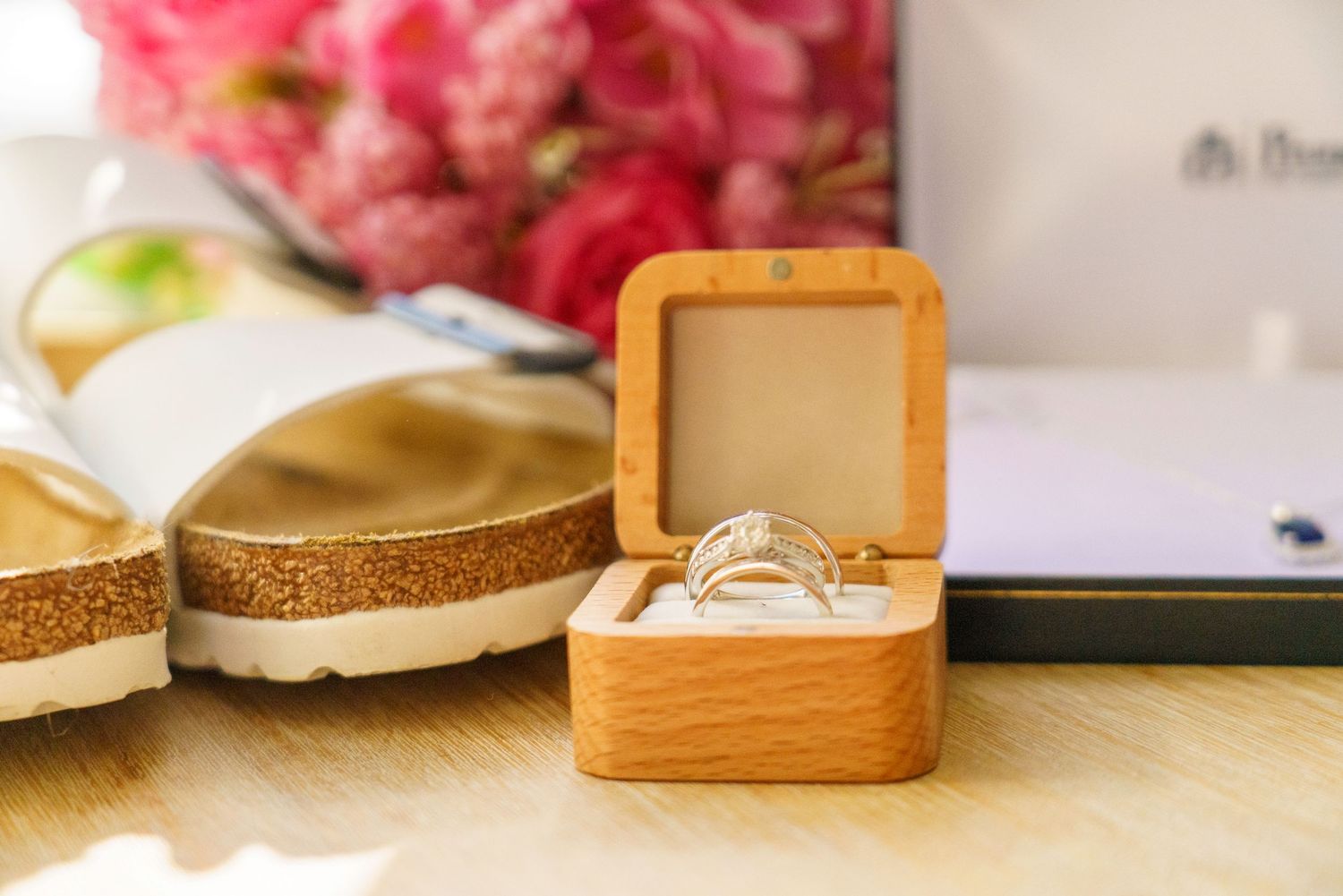 A diamond engagement ring displayed in an open wooden ring box on a table with sandals and pink flowers in the background.