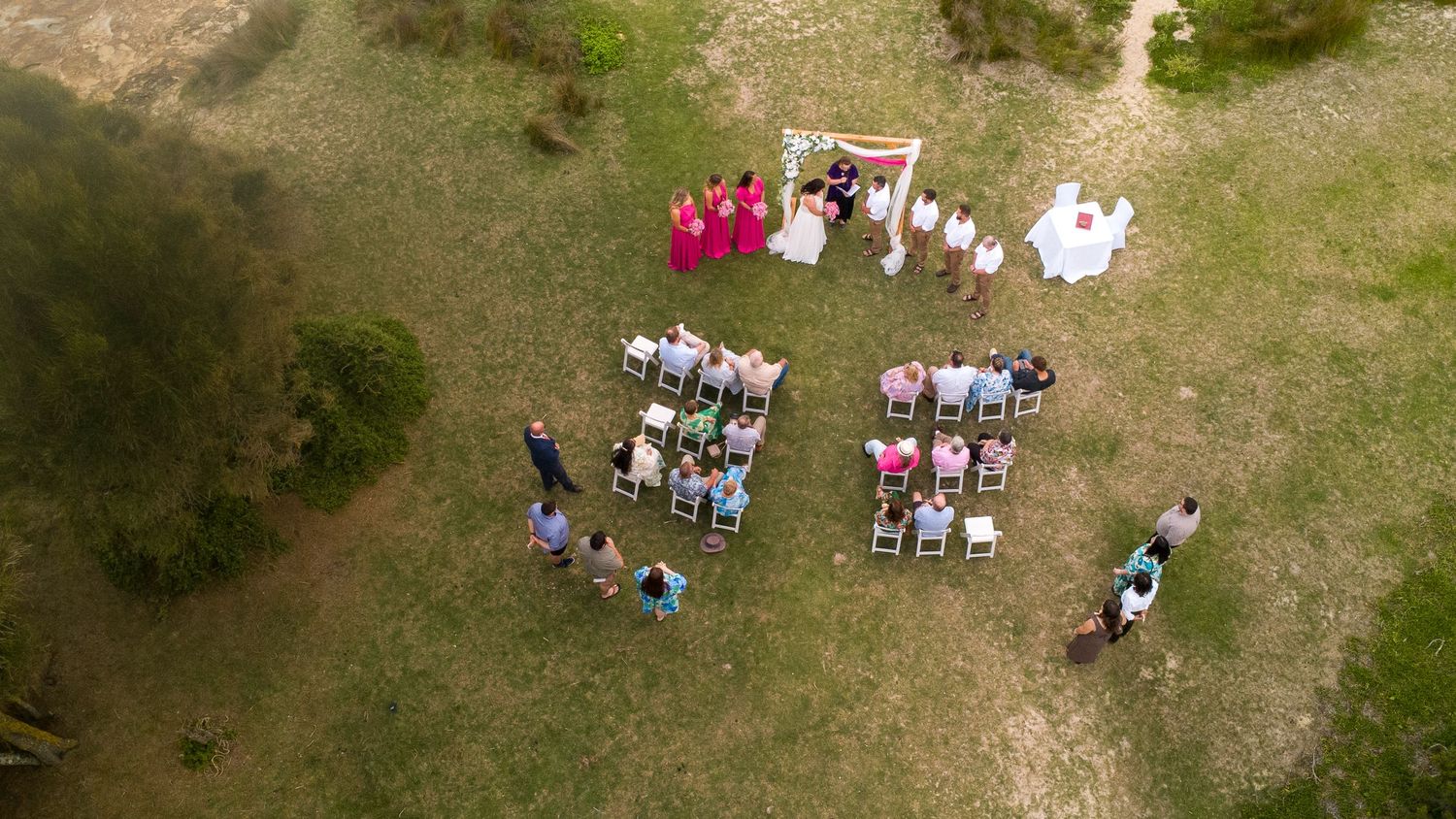 Aerial drone view of an outdoor wedding ceremony on green grass with guests seated in rows and bridal party.