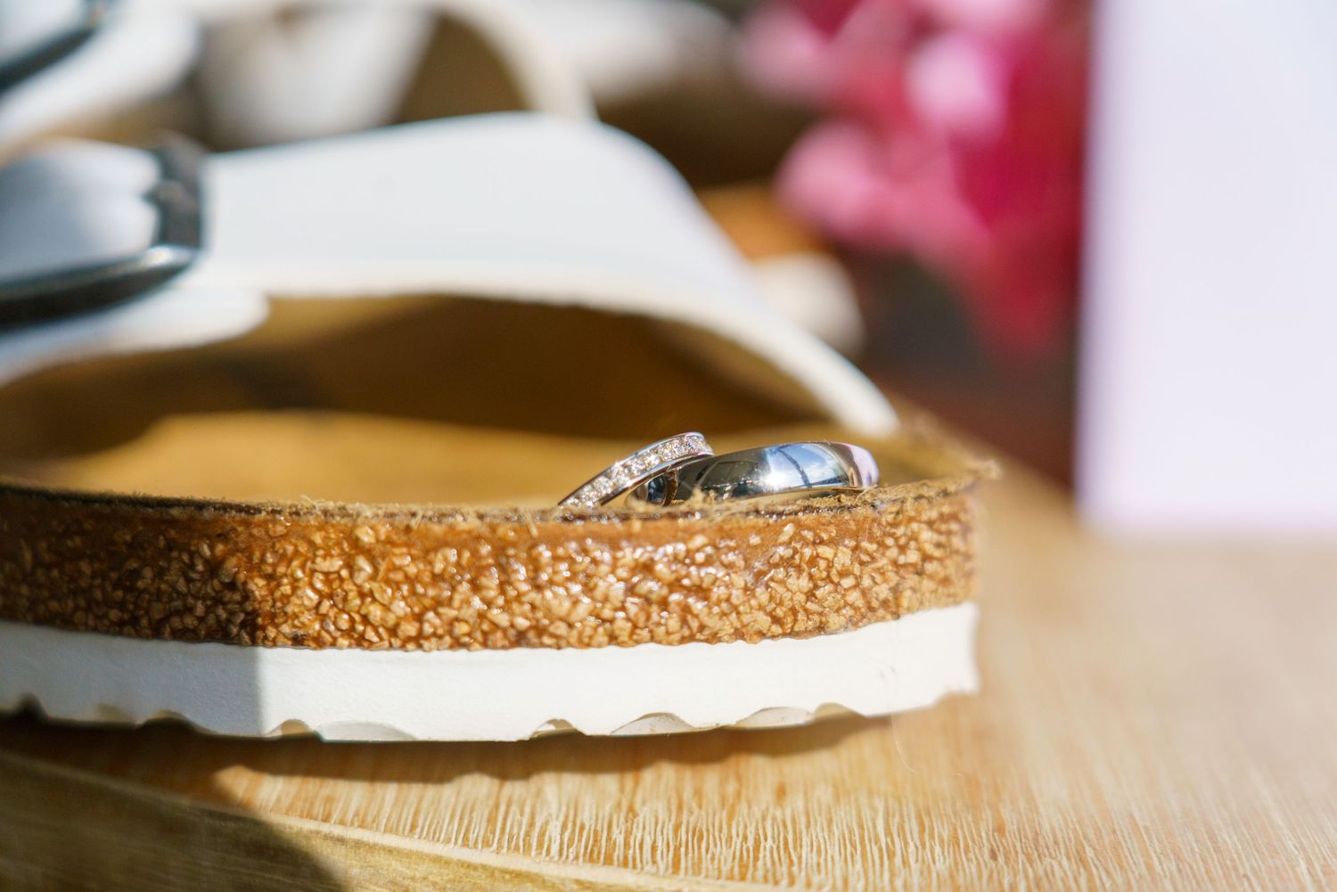 Wedding rings resting on a white cork-soled Birkenstock sandal on a wooden surface with soft floral bokeh background.
