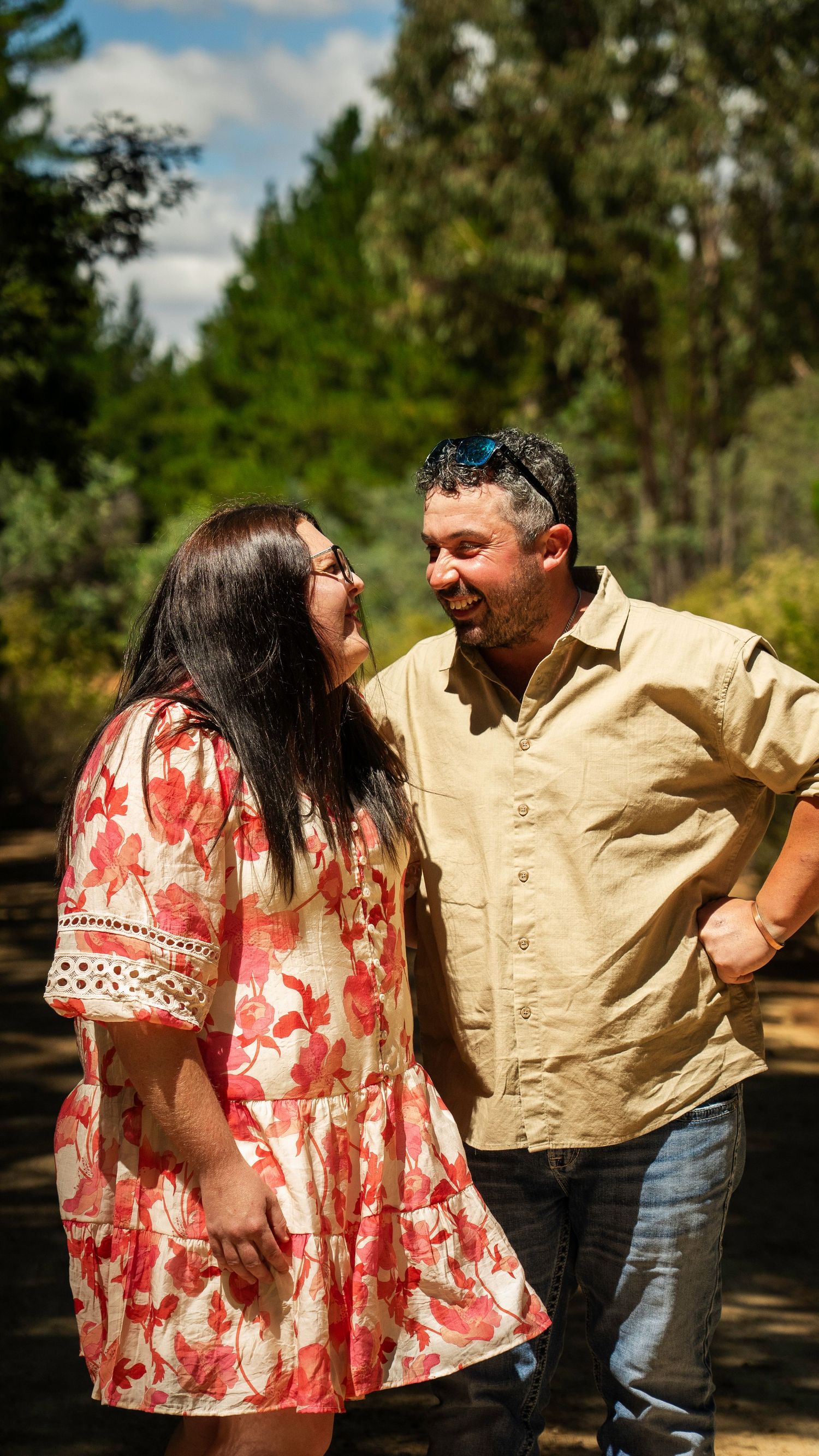 A smiling couple shares a joyful moment outdoors, woman in floral dress, man in beige shirt, surrounded by trees.