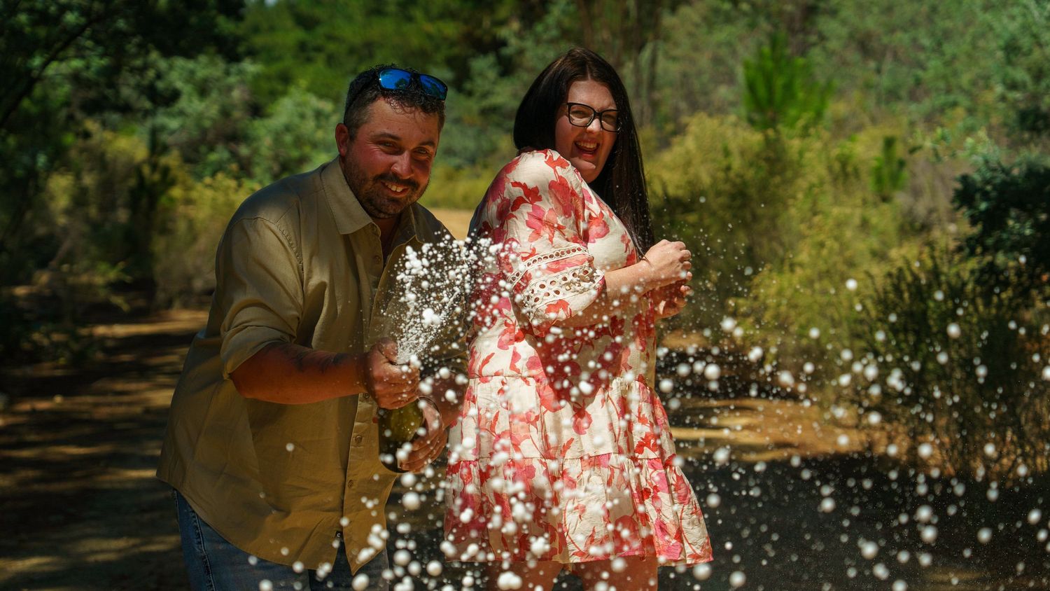 Couple joyfully splashing water outdoors surrounded by lush green trees on a sunny day.