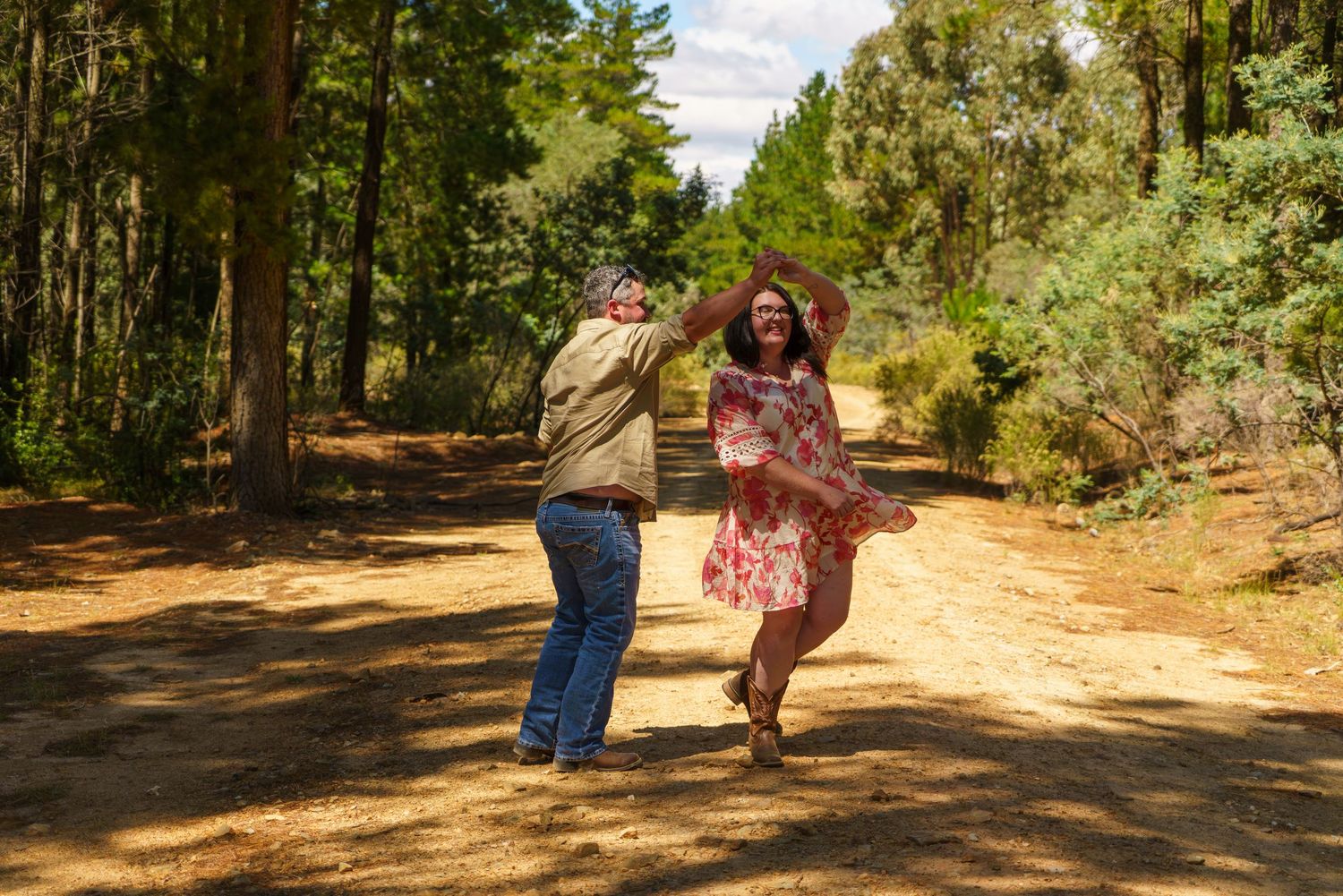 Couple dancing joyfully on a sunny dirt path surrounded by tall green trees in a forest setting.