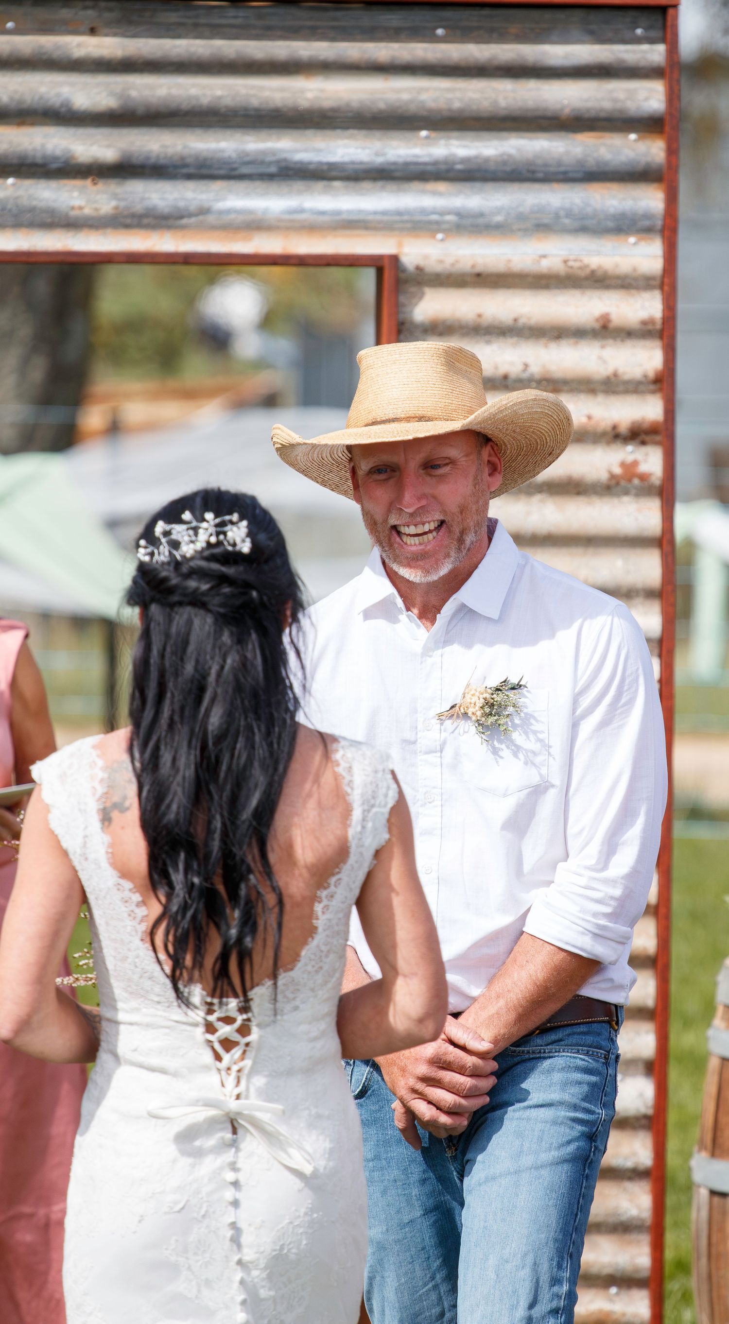 Smiling groom in cowboy hat and white shirt faces bride in lace wedding dress at rustic outdoor ceremony.