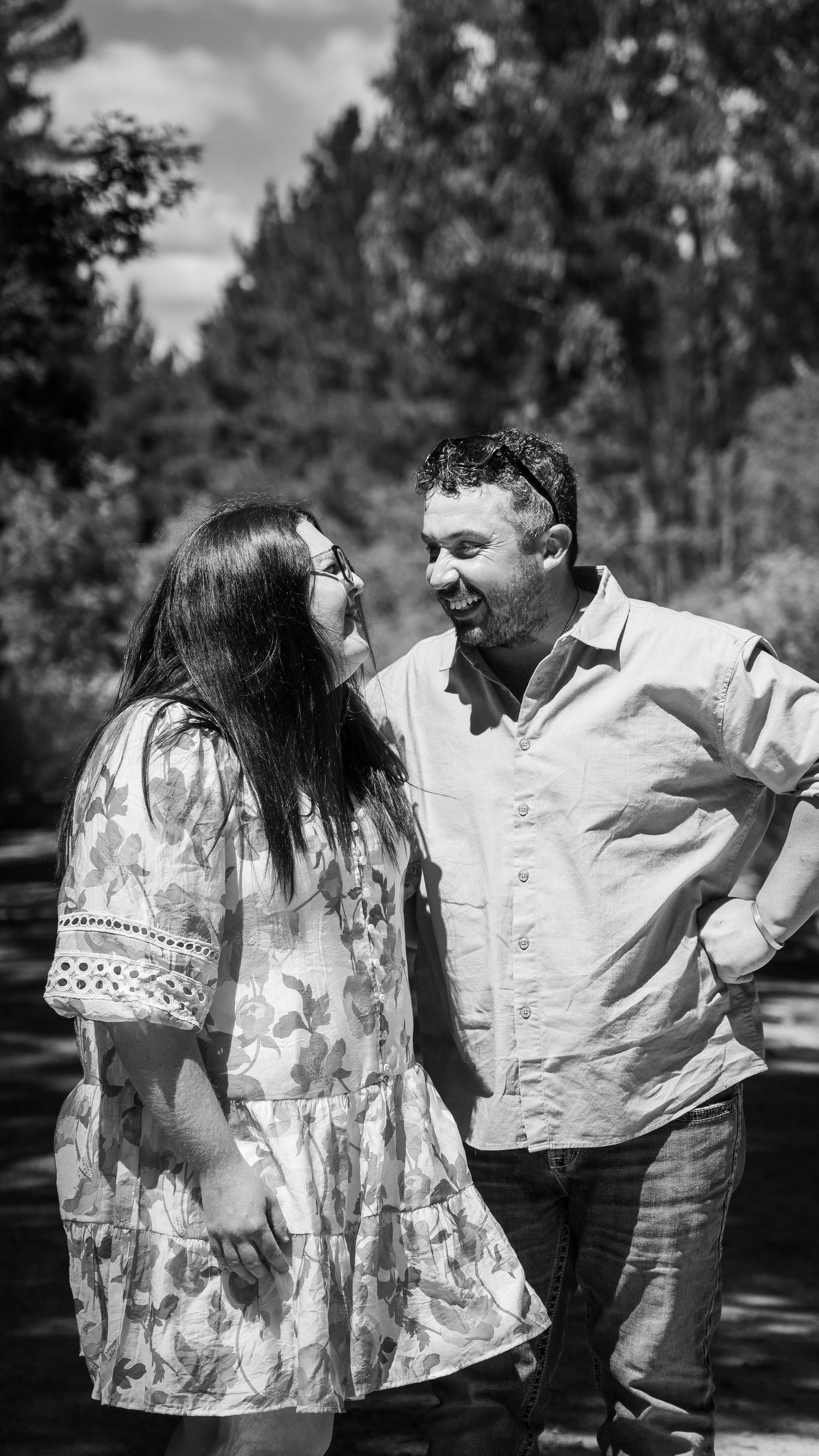 Black and white photo of a smiling couple standing outdoors surrounded by trees on a sunny day.