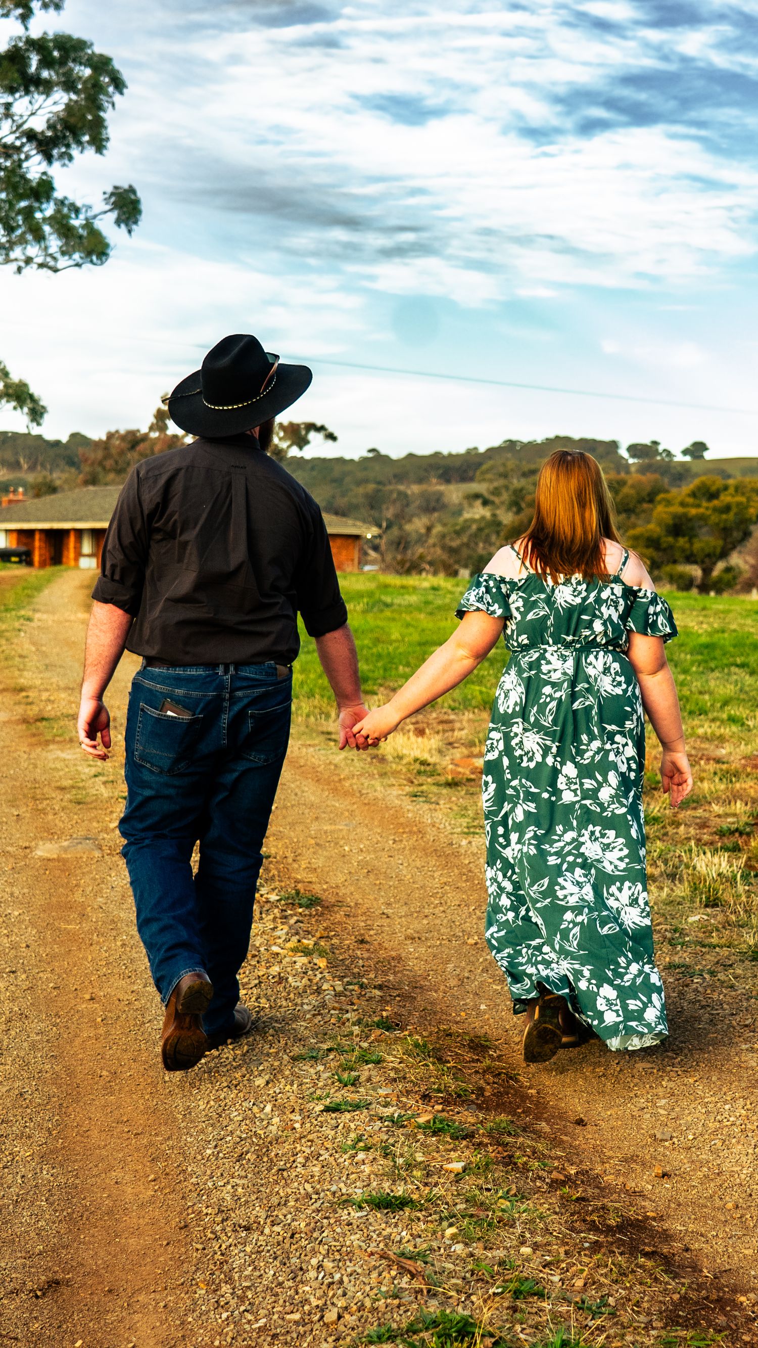 Couple in western attire walking hand in hand down rural dirt path.