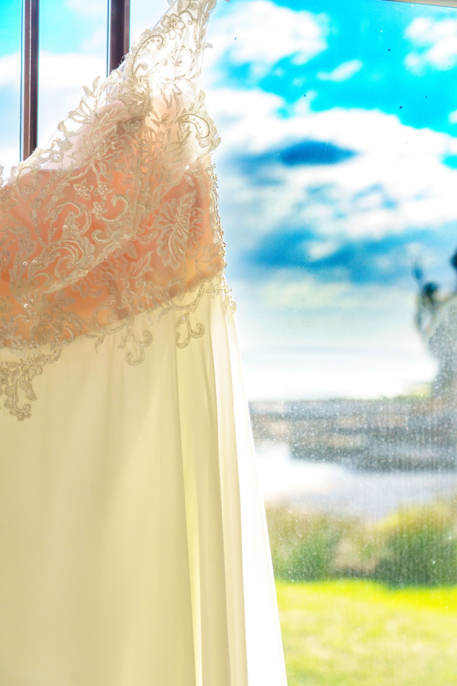 Elegant white wedding dress with lace bodice hanging by a window with blue sky and green lawn backdrop.