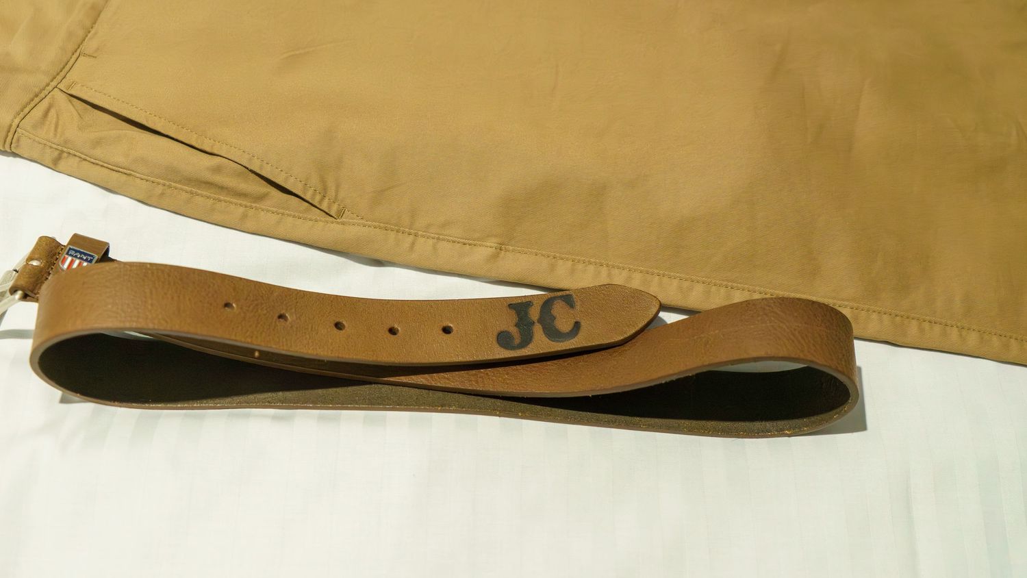 Brown leather belt with JC monogram initials displayed on khaki fabric background.