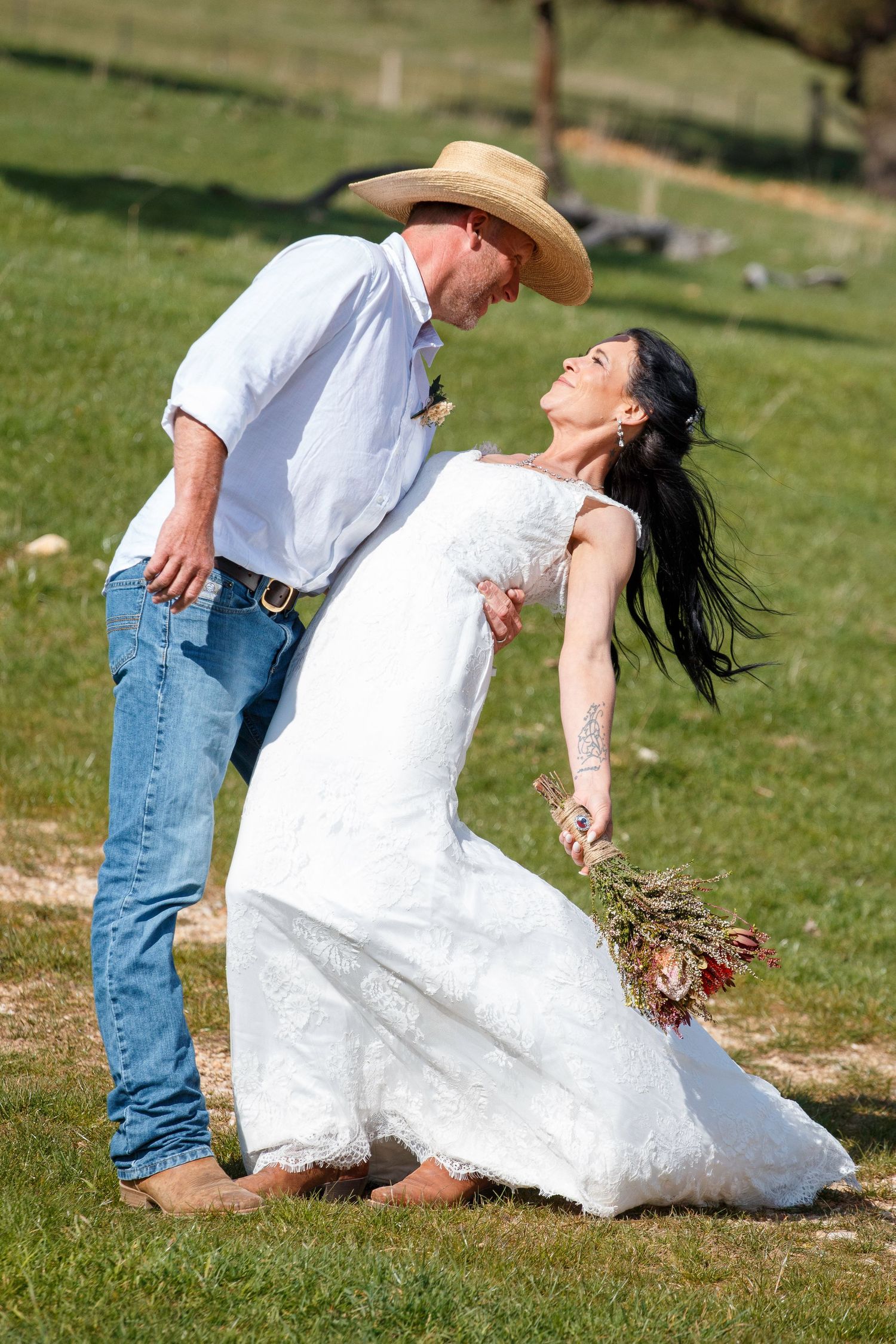 Bride in white lace gown holding bouquet dips with groom in cowboy hat and jeans during outdoor country wedding.