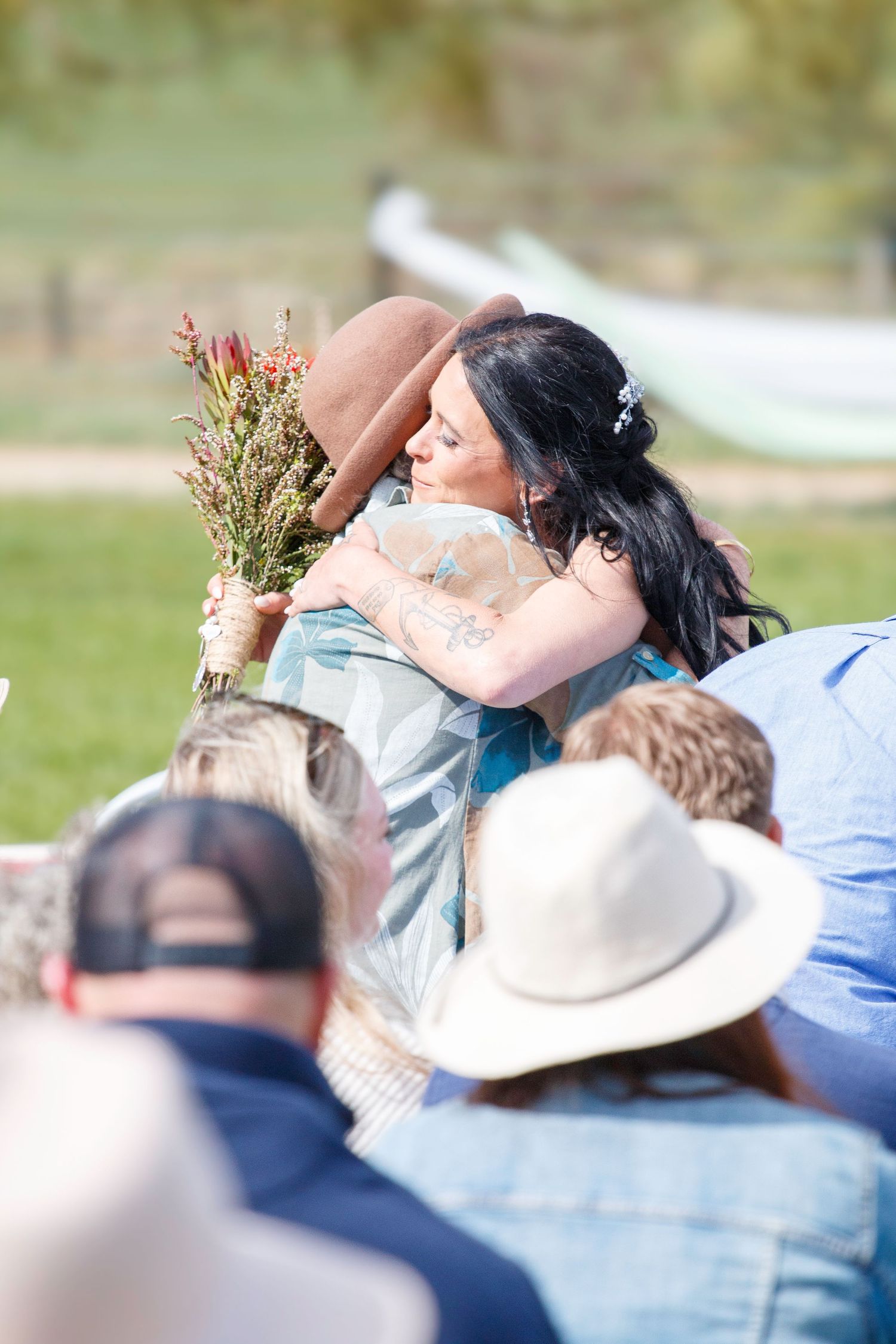 Bride with dark hair hugging a guest holding a wildflower bouquet at an outdoor wedding ceremony.