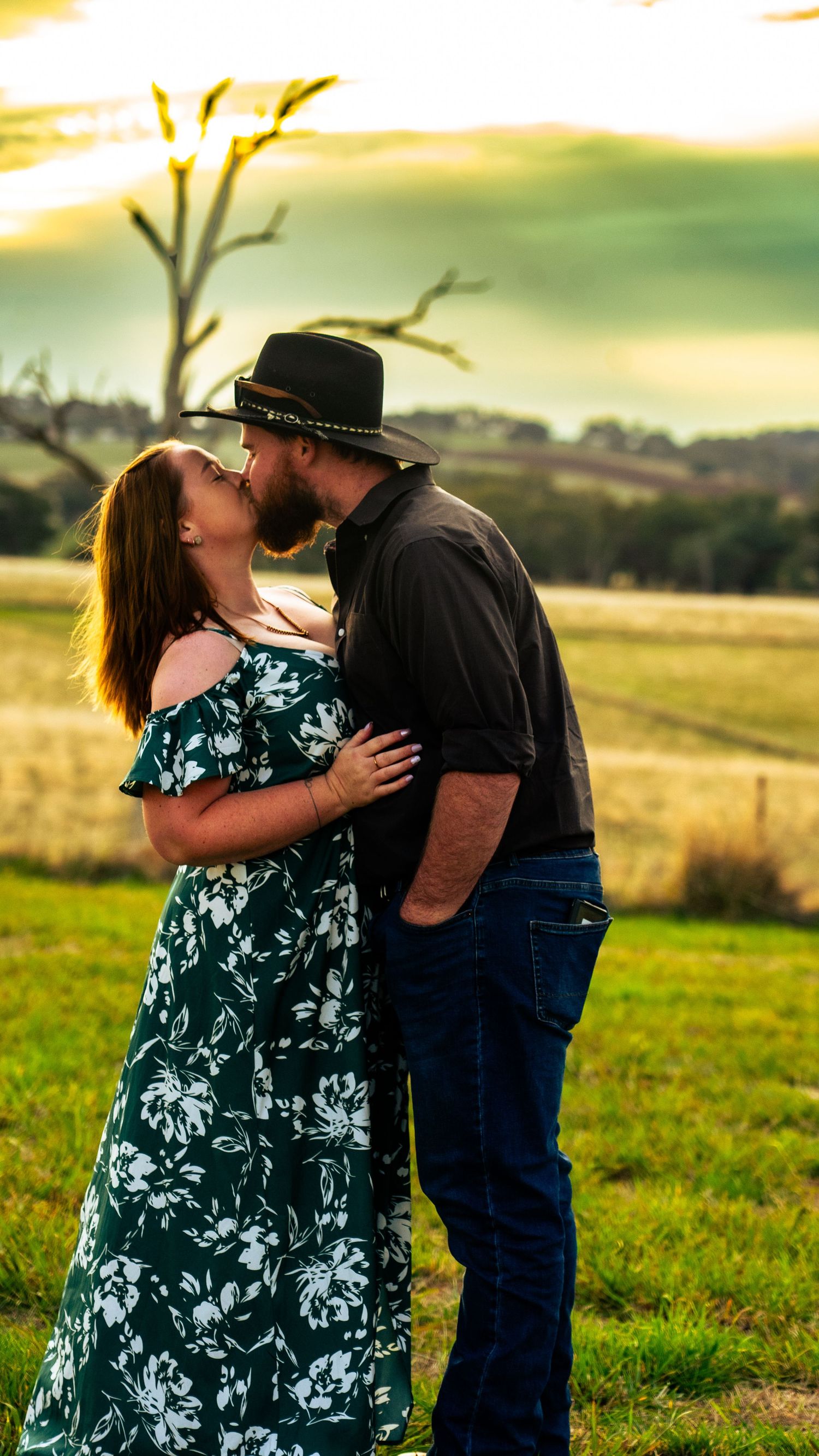 Romantic couple in country attire share tender moments at sunset in a rural field.