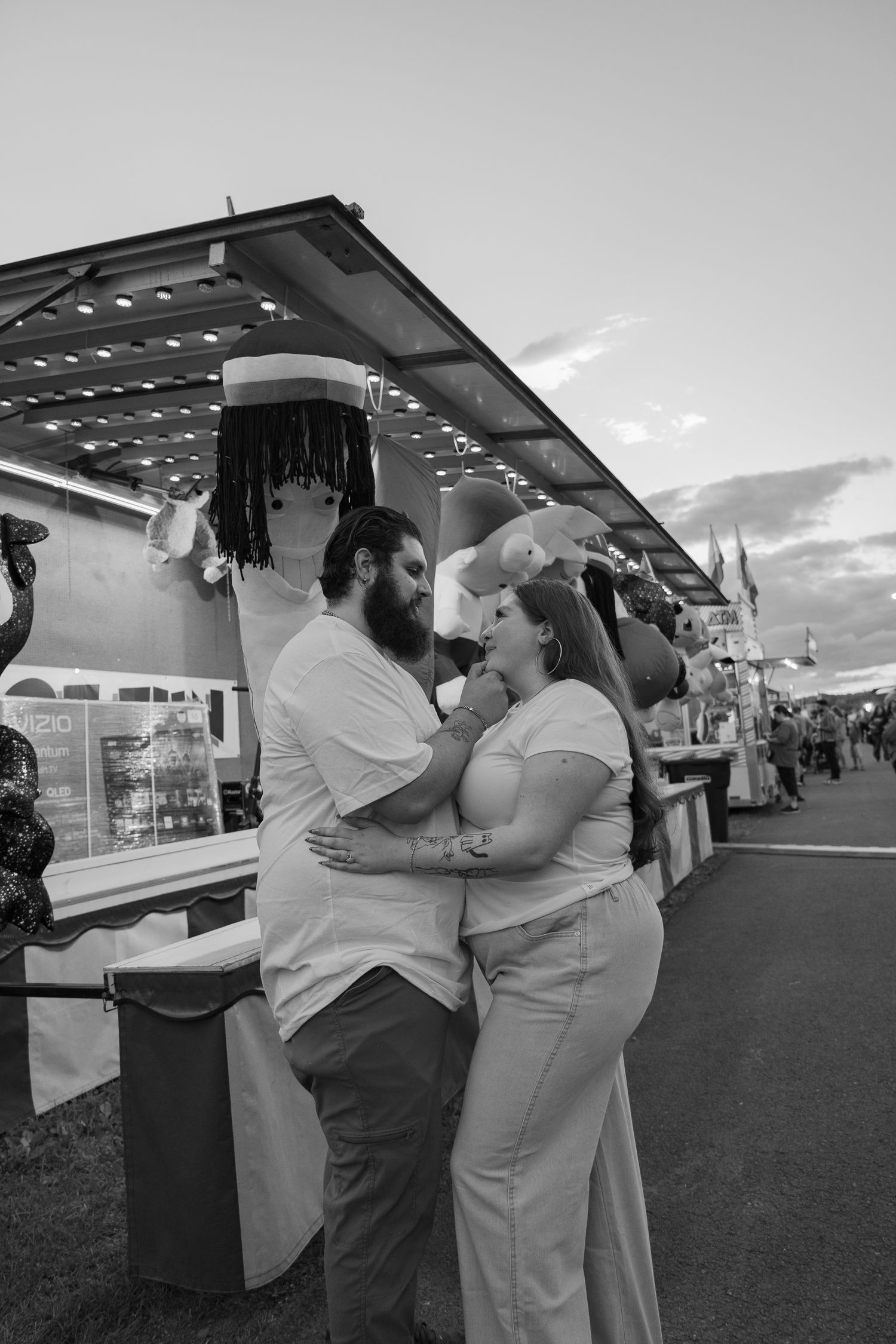 Black and white photo of a couple embracing at a fairground, with prize booths and stuffed animals in the background.