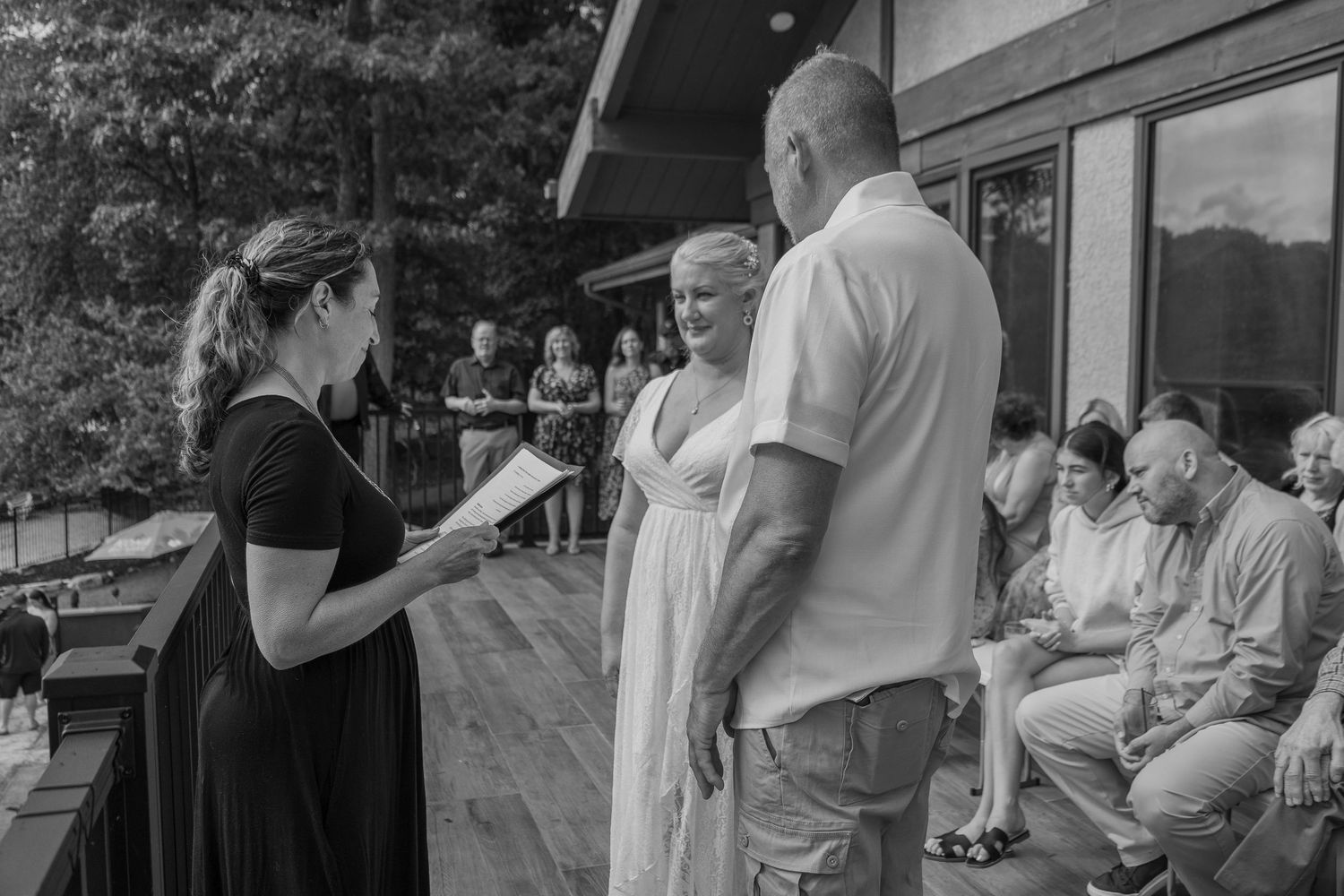 Outdoor wedding ceremony on wooden deck with officiant reading vows to couple before guests.