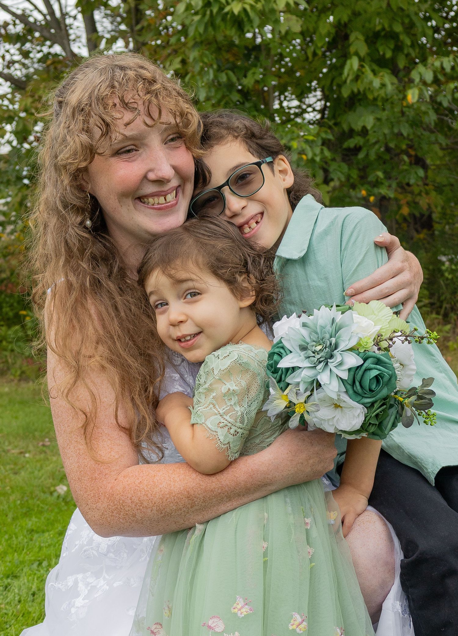 Woman with curly red hair hugs two children in mint green outfits outdoors, holding a floral bouquet.