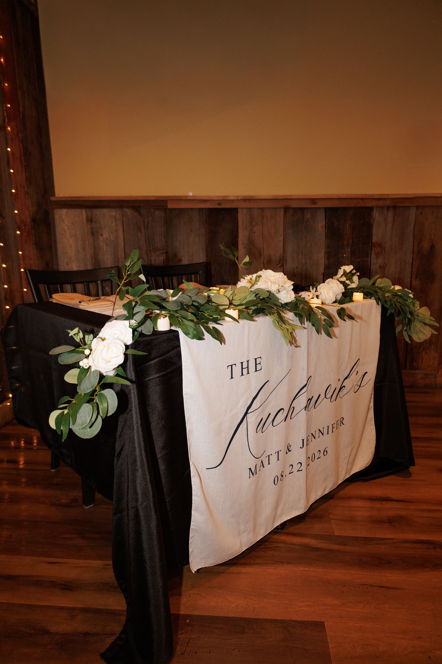 Elegant wedding sweetheart table with black linen, white floral garland, and personalized Kucharski name banner dated 08.22.2020.