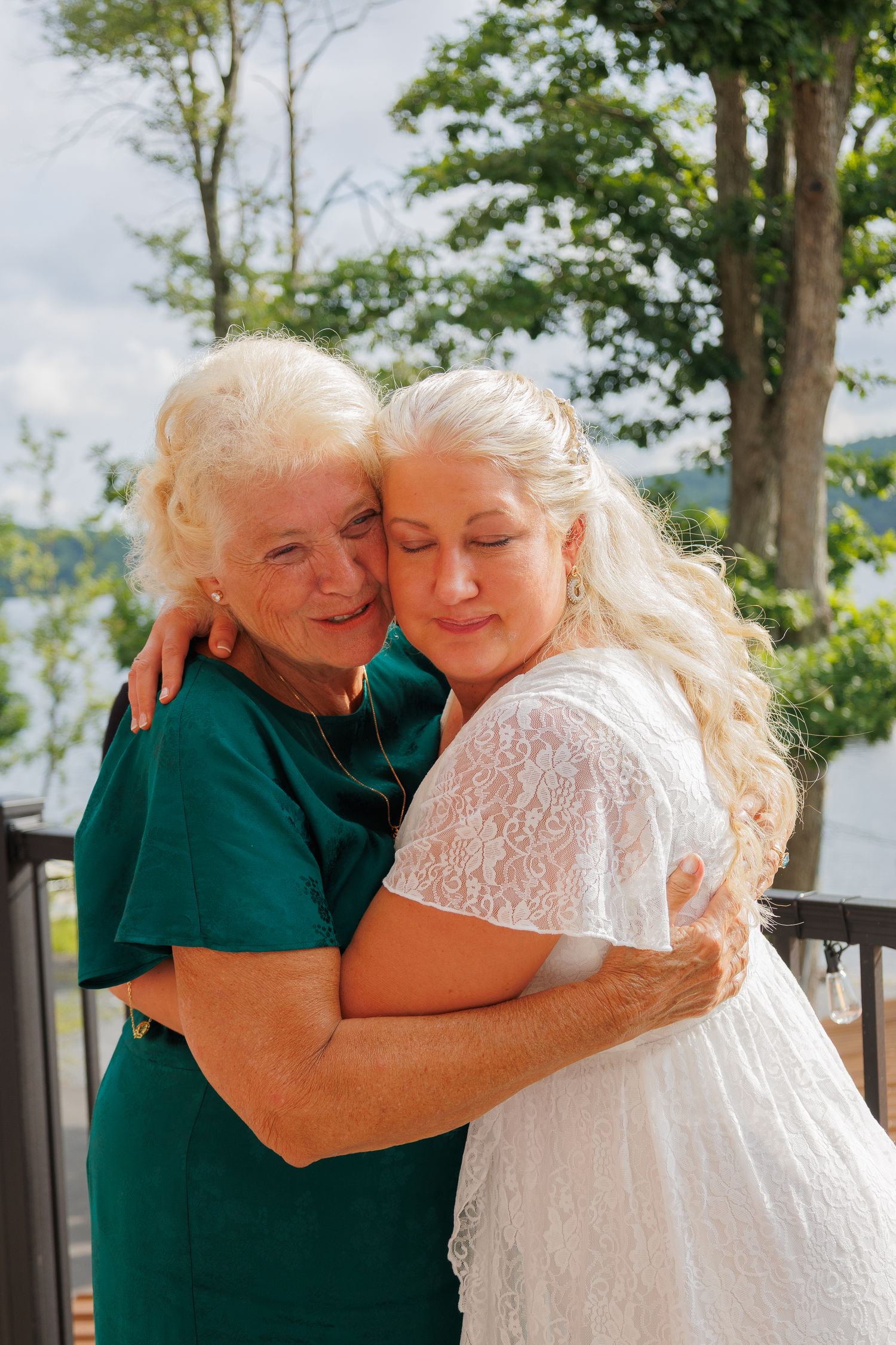 Bride in white lace dress embracing elderly woman in teal dress outdoors at wedding.