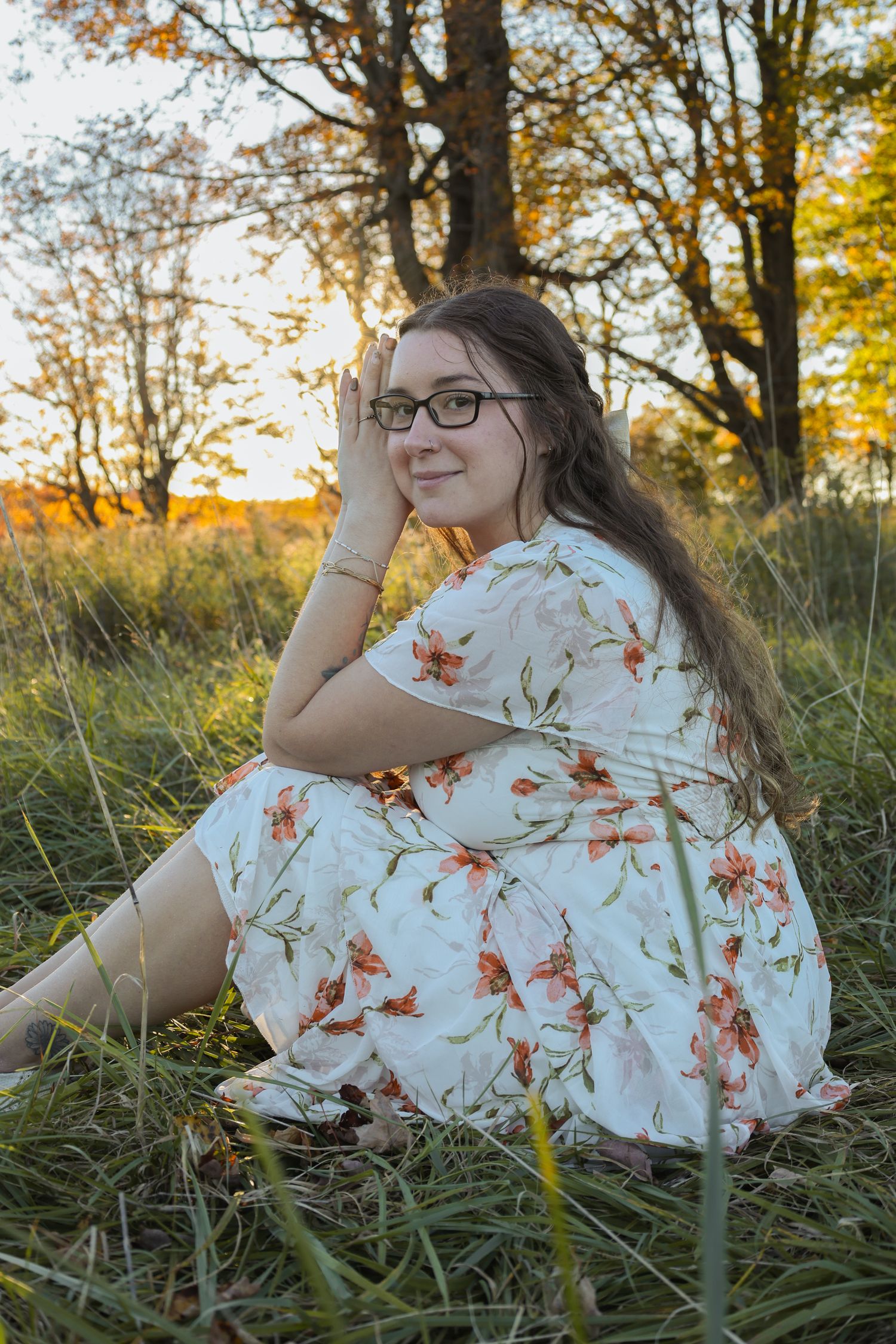 Young woman in floral dress sitting in grass at golden hour sunset, smiling with glasses and long hair.