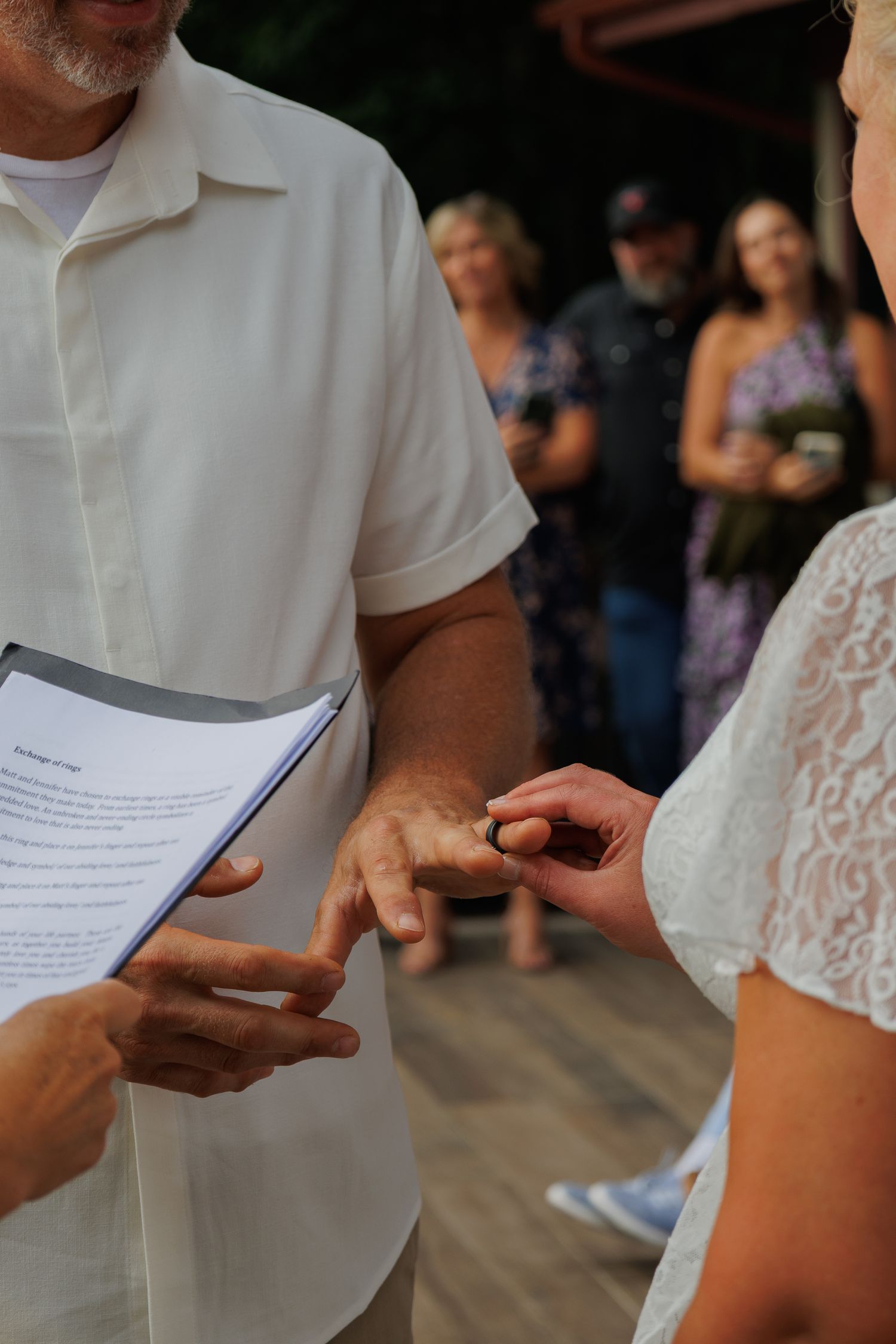Couple exchanging wedding rings during an outdoor ceremony while officiant holds printed vows.