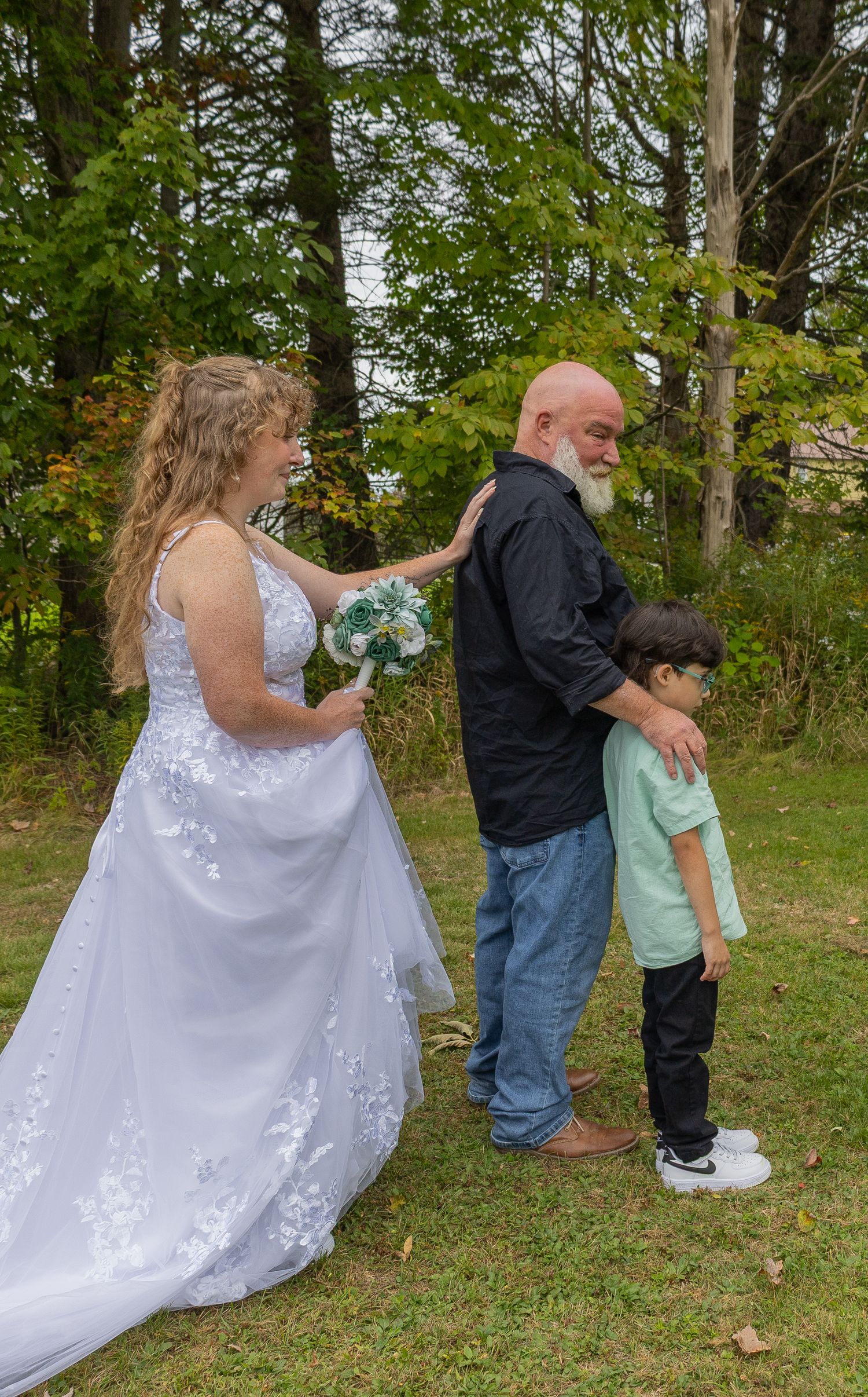 Bride in white gown holds bouquet beside bald groom and young boy during outdoor wedding ceremony near trees.
