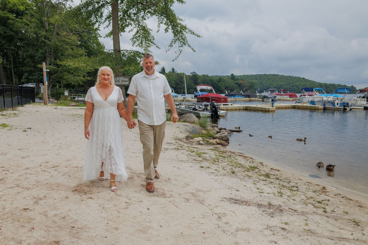 Couple in wedding attire walking hand in hand on sandy lakeside beach with marina in background.