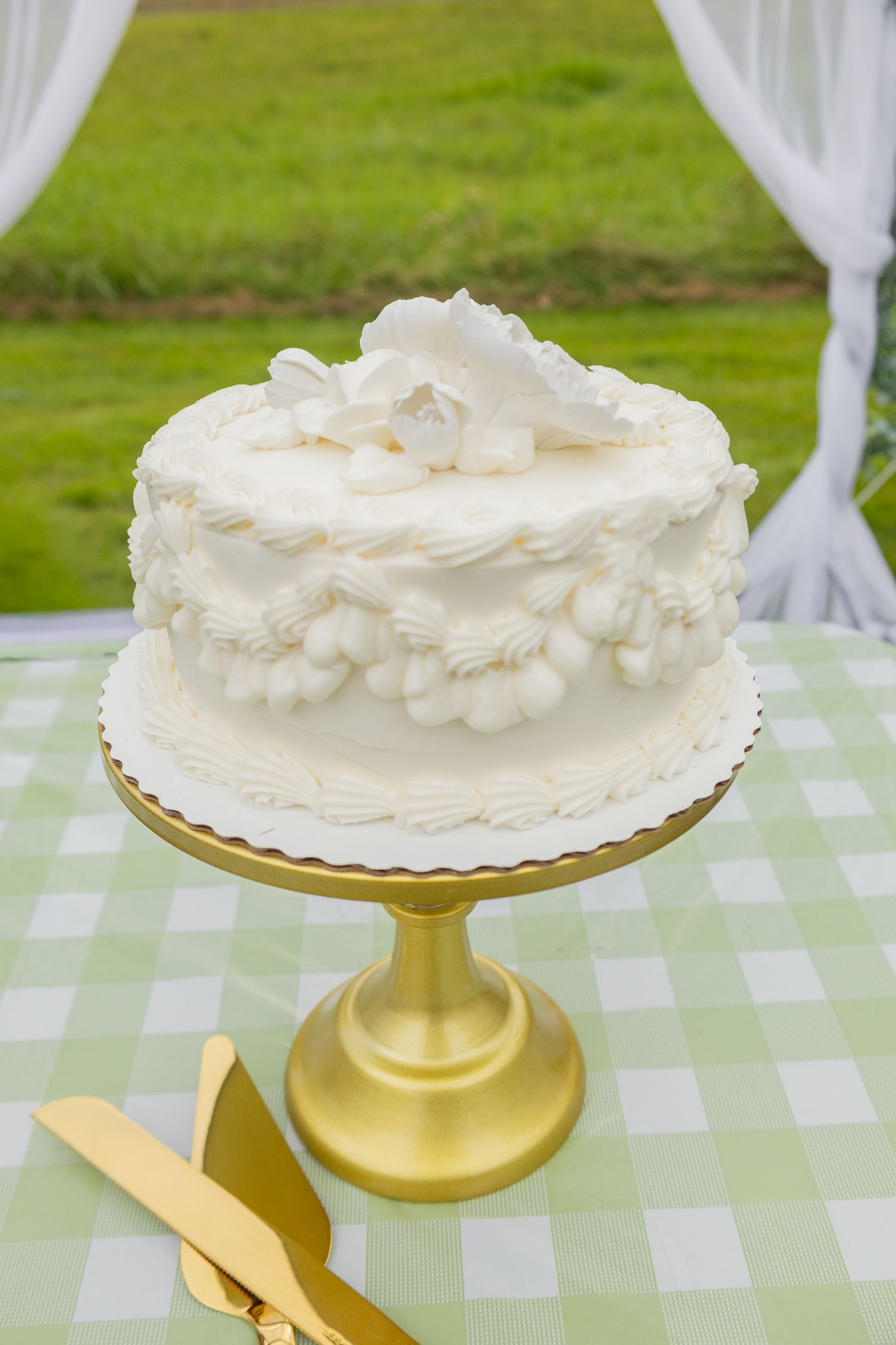 Elegant white wedding cake with floral frosting on a gold pedestal stand, displayed on a green gingham tablecloth outdoors.