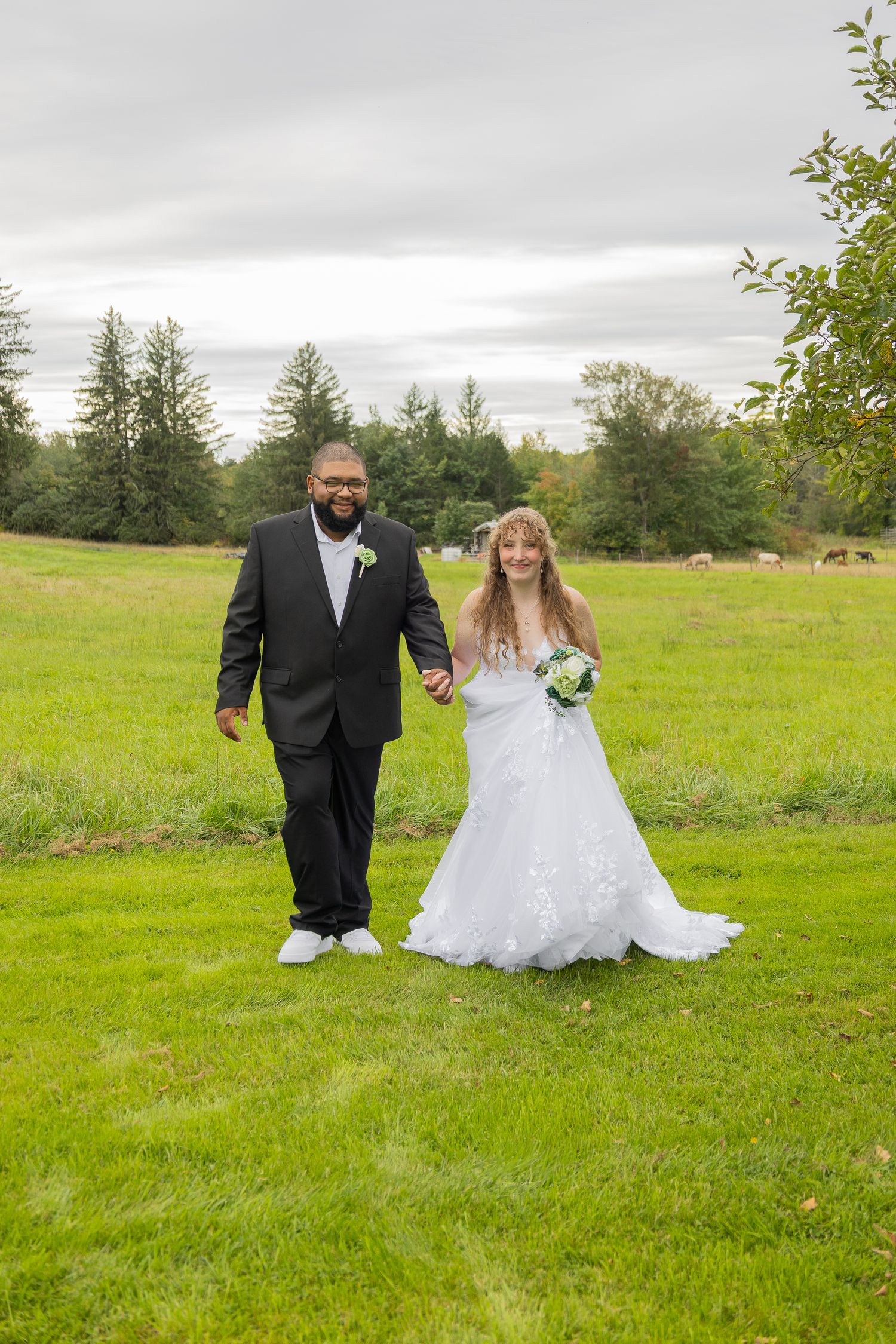 Newlywed couple holding hands walking on green grass, bride in white gown with bouquet, groom in black suit, outdoor wedding.