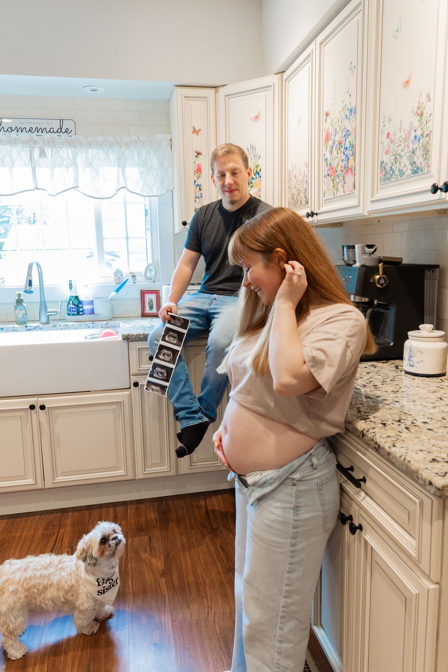 Pregnant woman and man holding ultrasound photos in a cozy kitchen with a small white dog wearing a bandana.