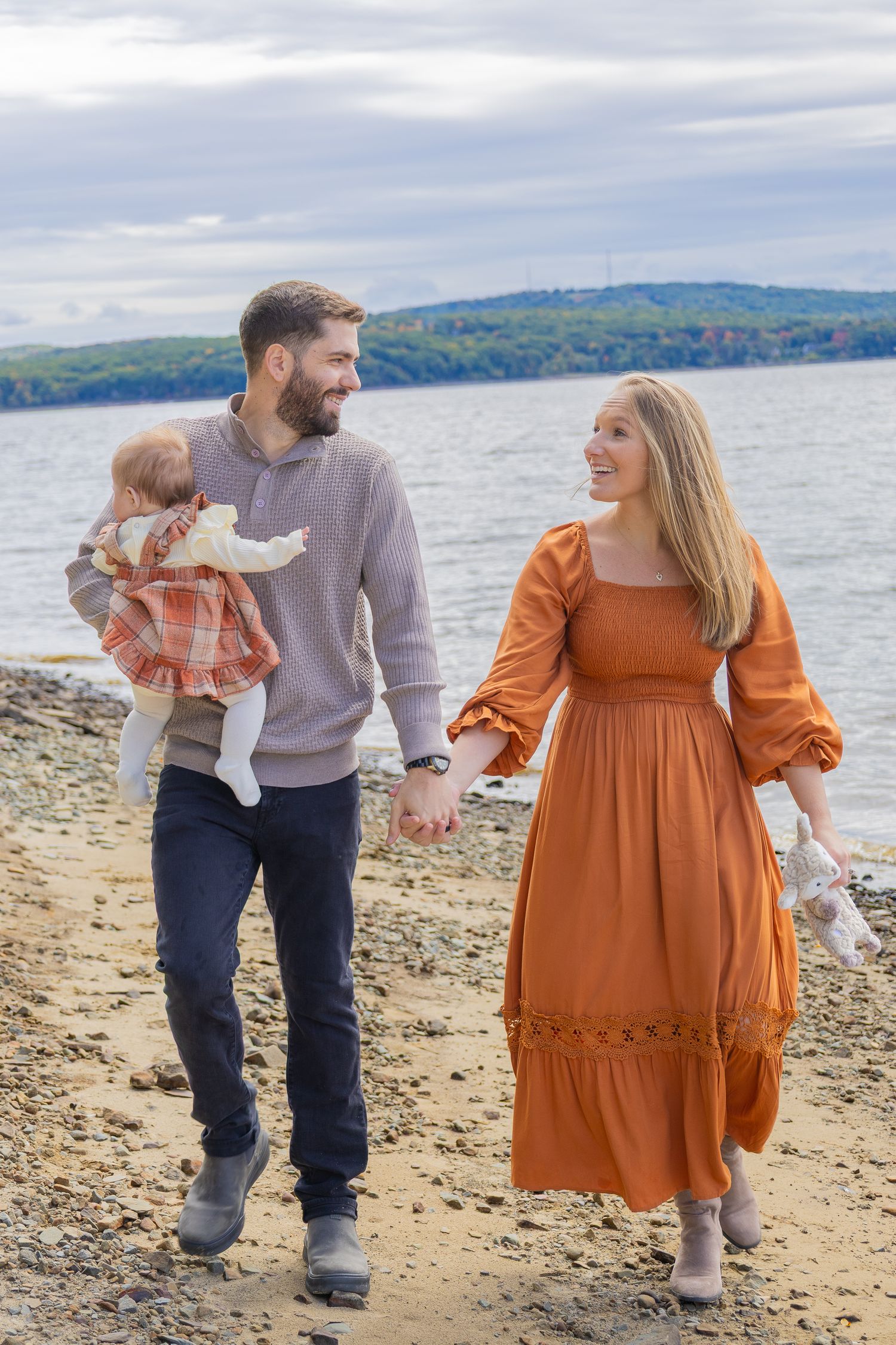 A young family walks along a rocky beach, man holding baby in plaid dress, woman in orange gown, hills in background.
